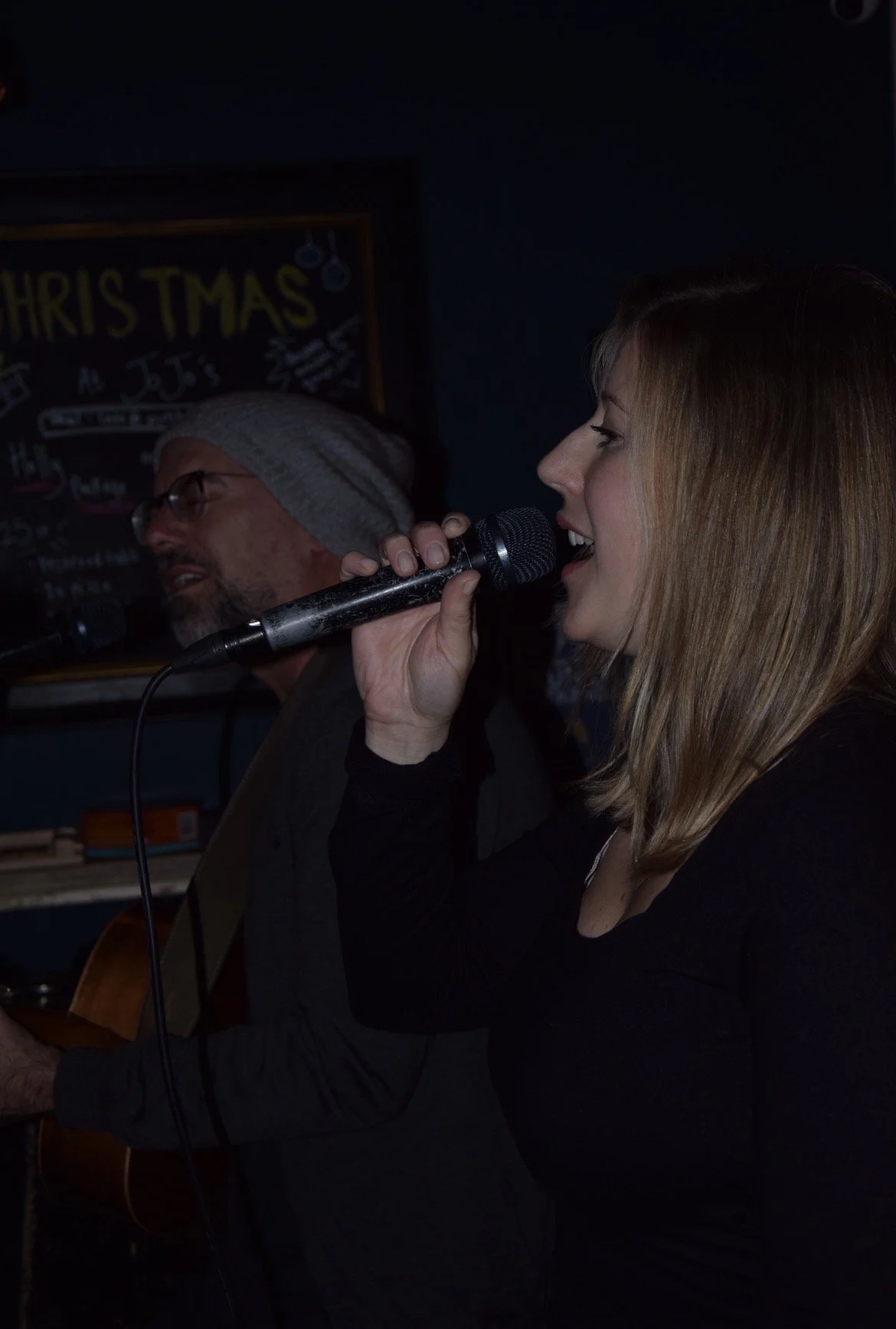 A woman singing into a microphone with a man playing guitar in the background, at JoJo's Open Mic Night. 