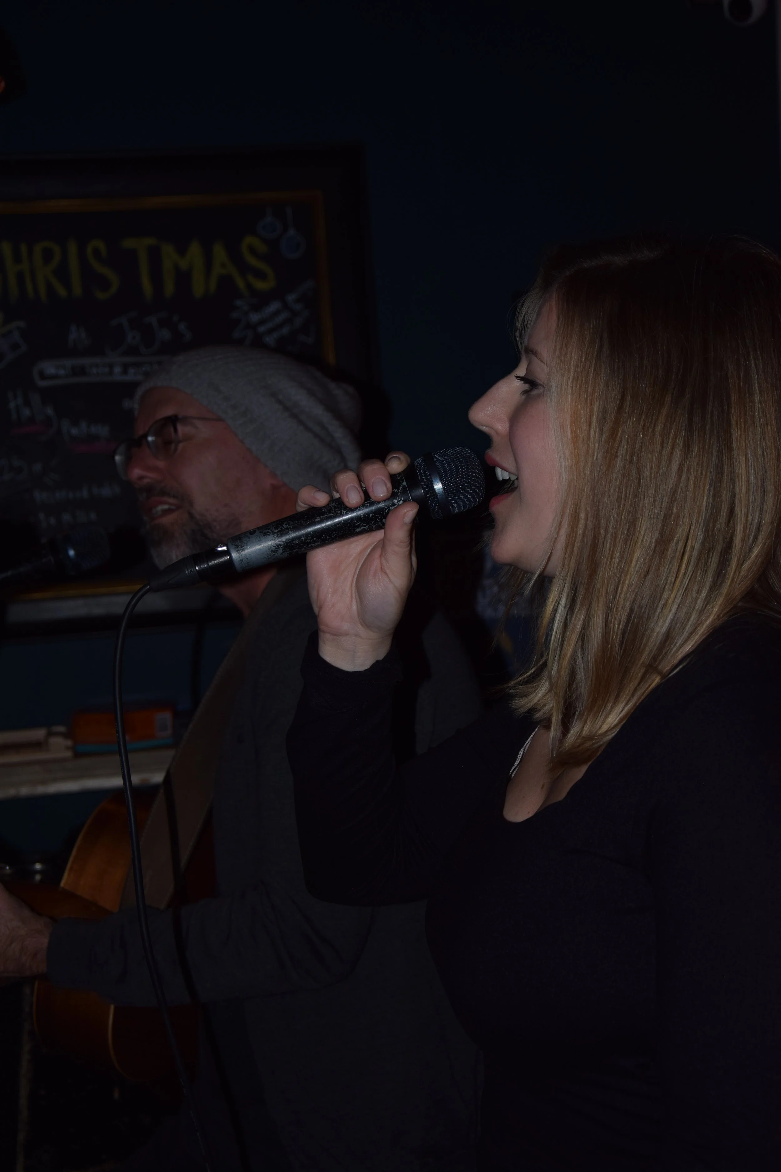 A woman singing into a microphone with a man playing guitar in the background, at JoJo's Open Mic Night. 