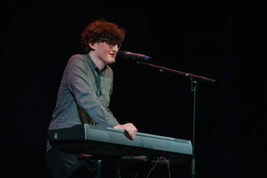 Jack McMinn playing a keyboard and singing a comedy song into a microphone on stage.