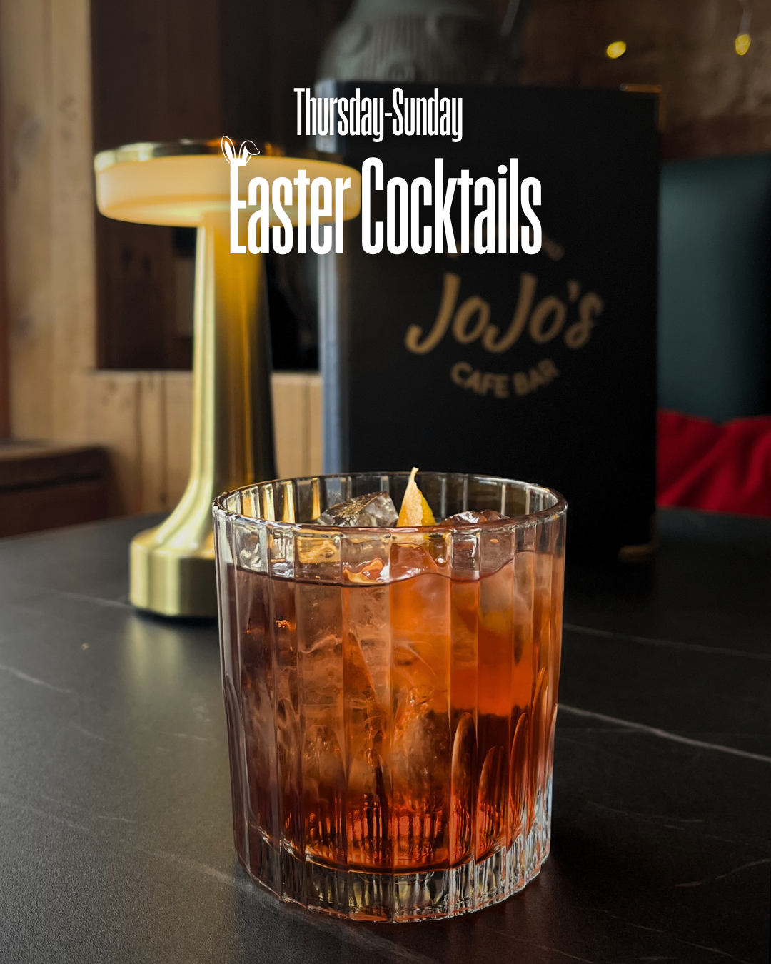 A glass of cocktail with ice and a lemon peel on a dark table, with a black menu stand with gold lettering and a modern table lamp in the background, promoting Easter cocktails available Thursday through Sunday at JoJo's Cocktail & Pizza Bar, Oxford.