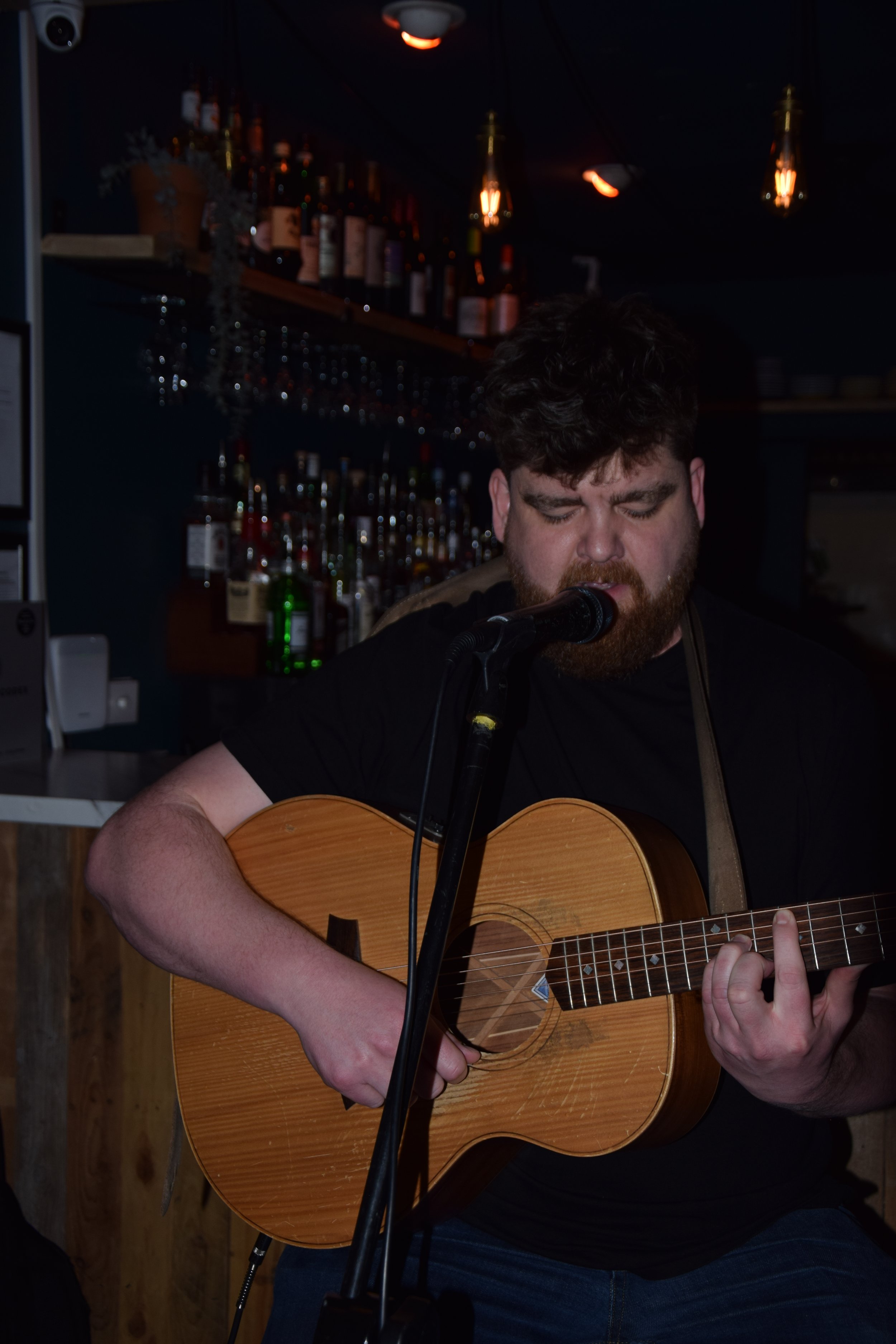 A man intensely sings into a microphone while playing an acoustic guitar at the Open Mic Night at JoJo's Cocktail bar, Oxford, with shelves of liquor bottles in the background.