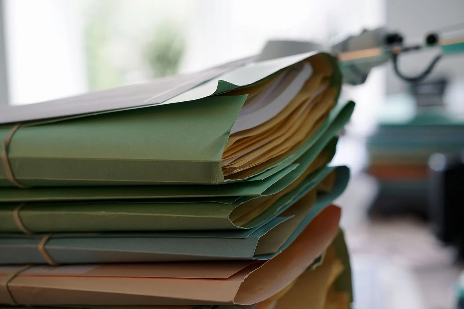 Høj stak papirfiler bundet sammen, placeret på en bordflade, med en baggrund af et kontormiljø.