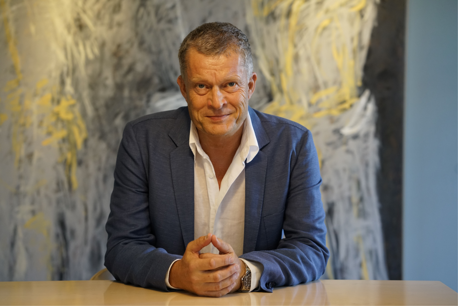 Henrik Hougaard sidder ved et bord iført mørk blazer og hvid skjorte, med kunstnerisk maleri i baggrunden.