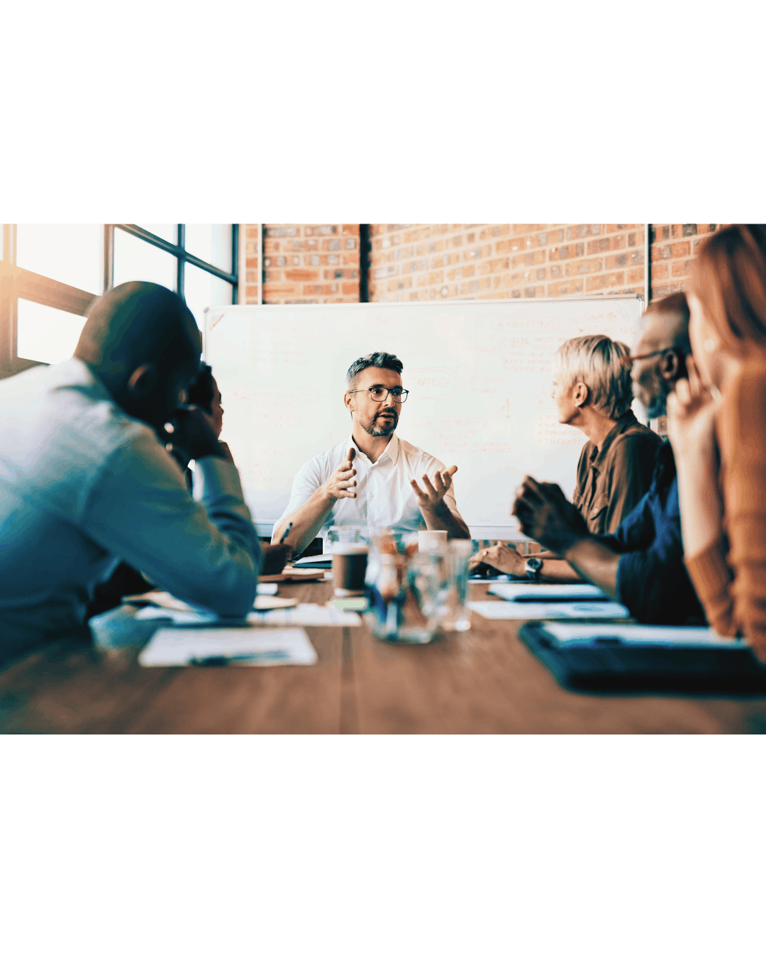 A man is leading a meeting with five people seated around a table, with a whiteboard behind him in a brick-walled room.