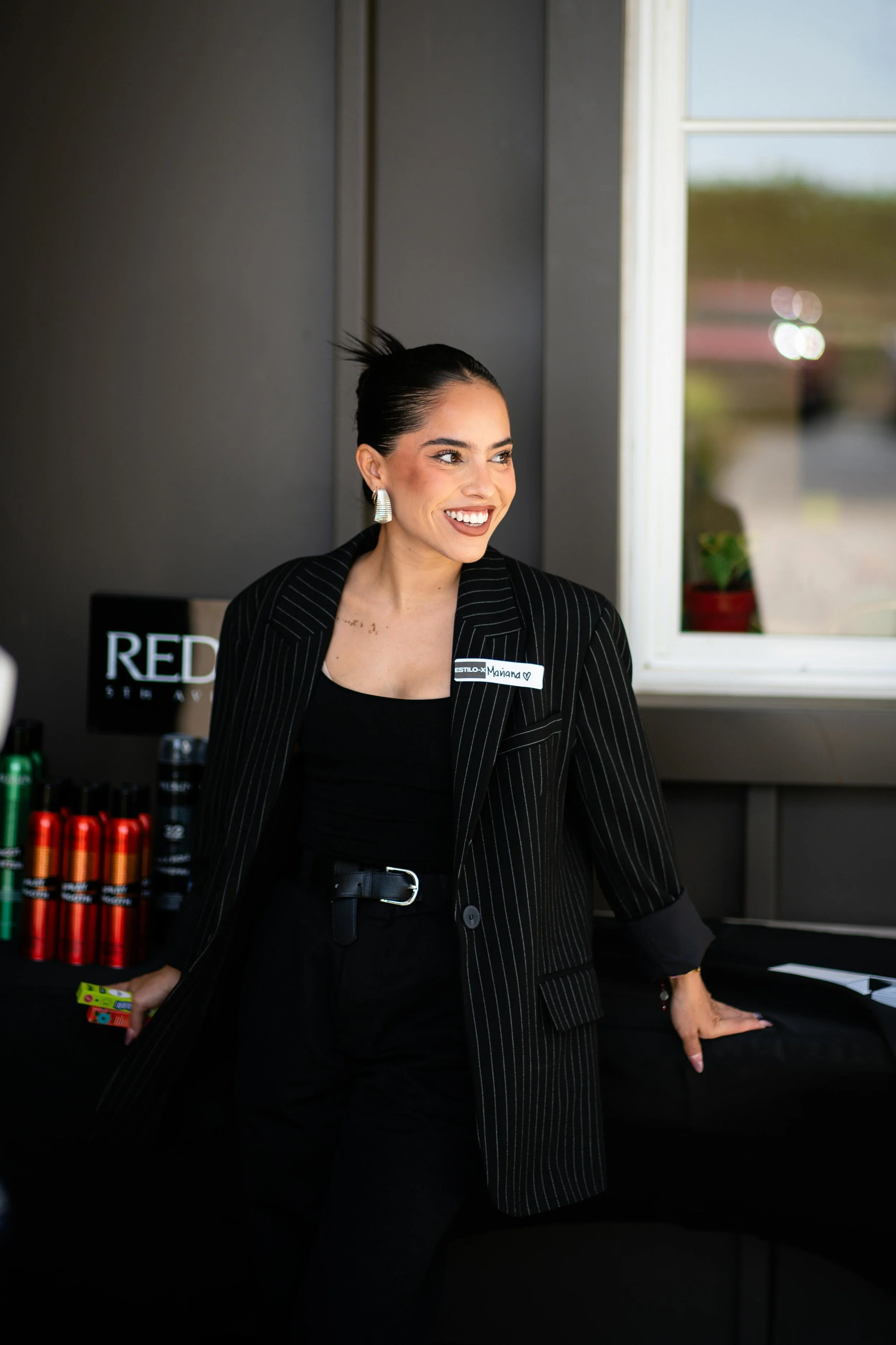 A woman with dark hair pulled back, wearing a black pinstripe blazer and black top, smiling and standing at a table with various colored bottles.