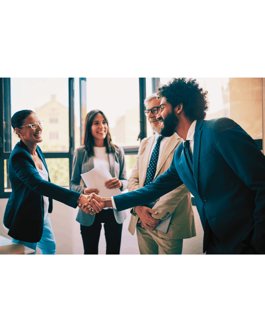 Business professionals shaking hands in an office, smiling, with three other colleagues observing in the background.