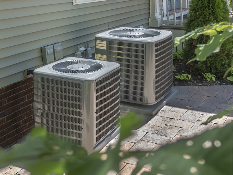 Two outdoor HVAC units next to a house wall and patio with plants in the foreground.