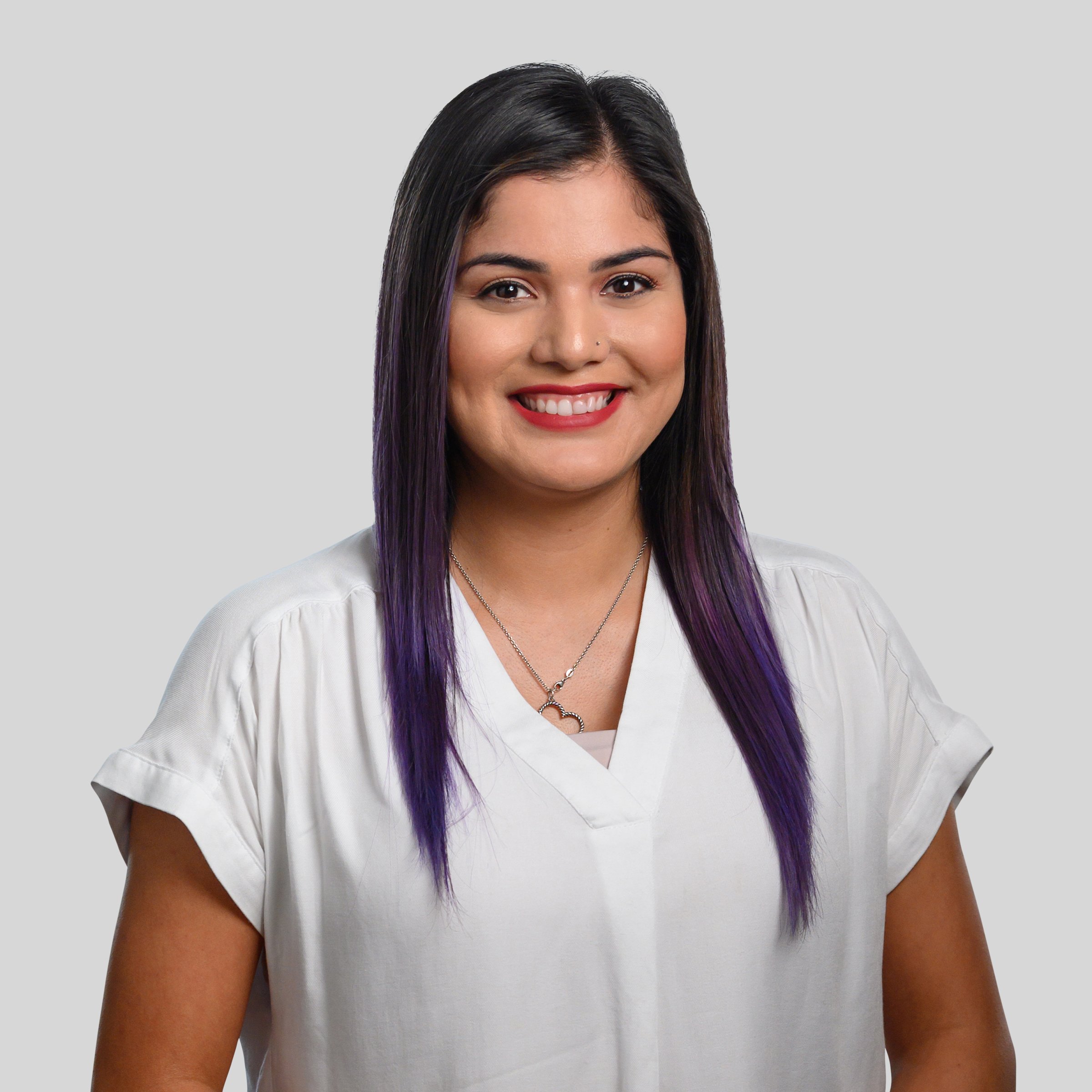 A woman with long black hair, purple tips, and a nose ring, smiling in studio against a plain light gray background, wearing a white top and a silver necklace.