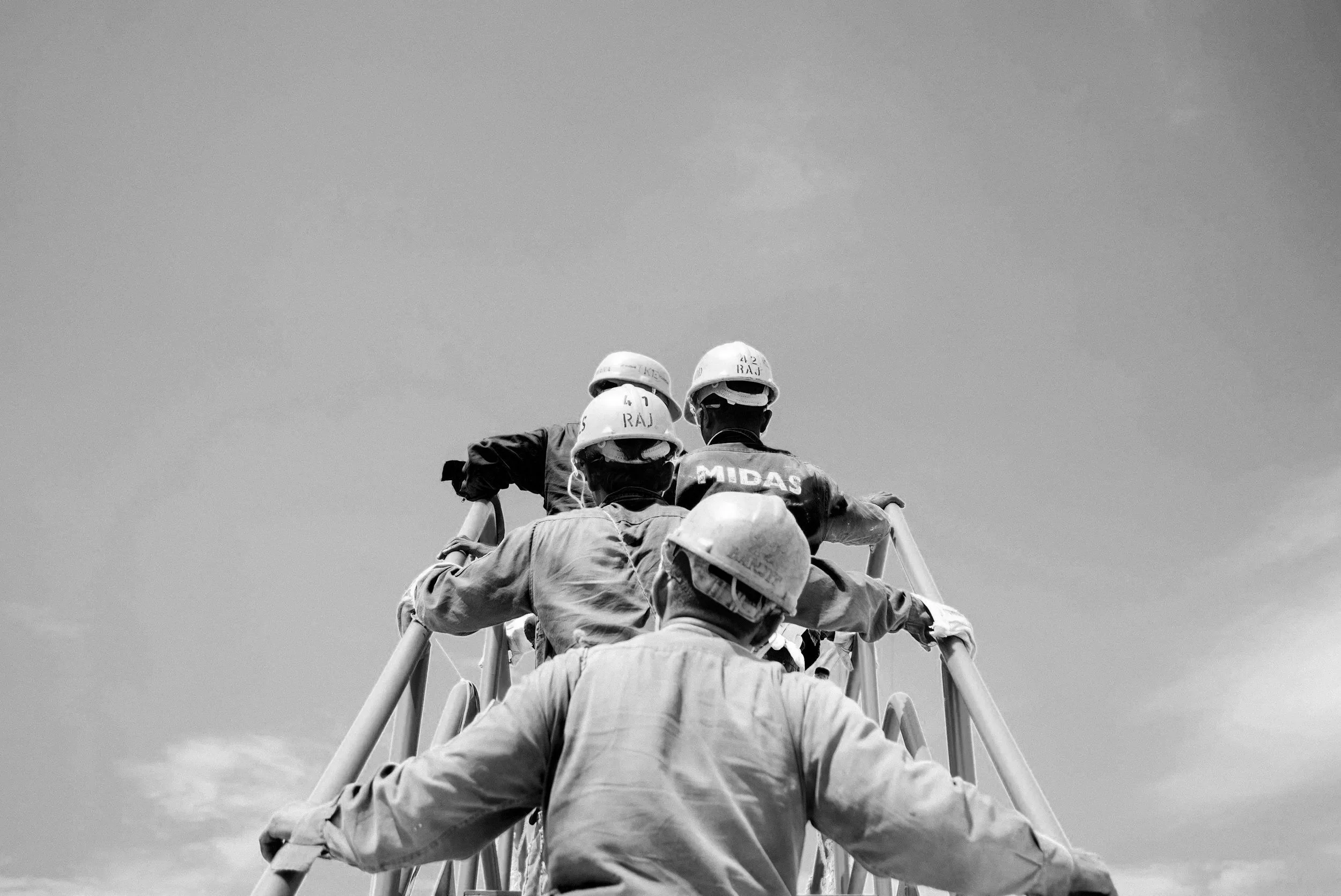 Group of workers wearing safety helmets climbing a metal ladder outdoors under a cloudy sky.