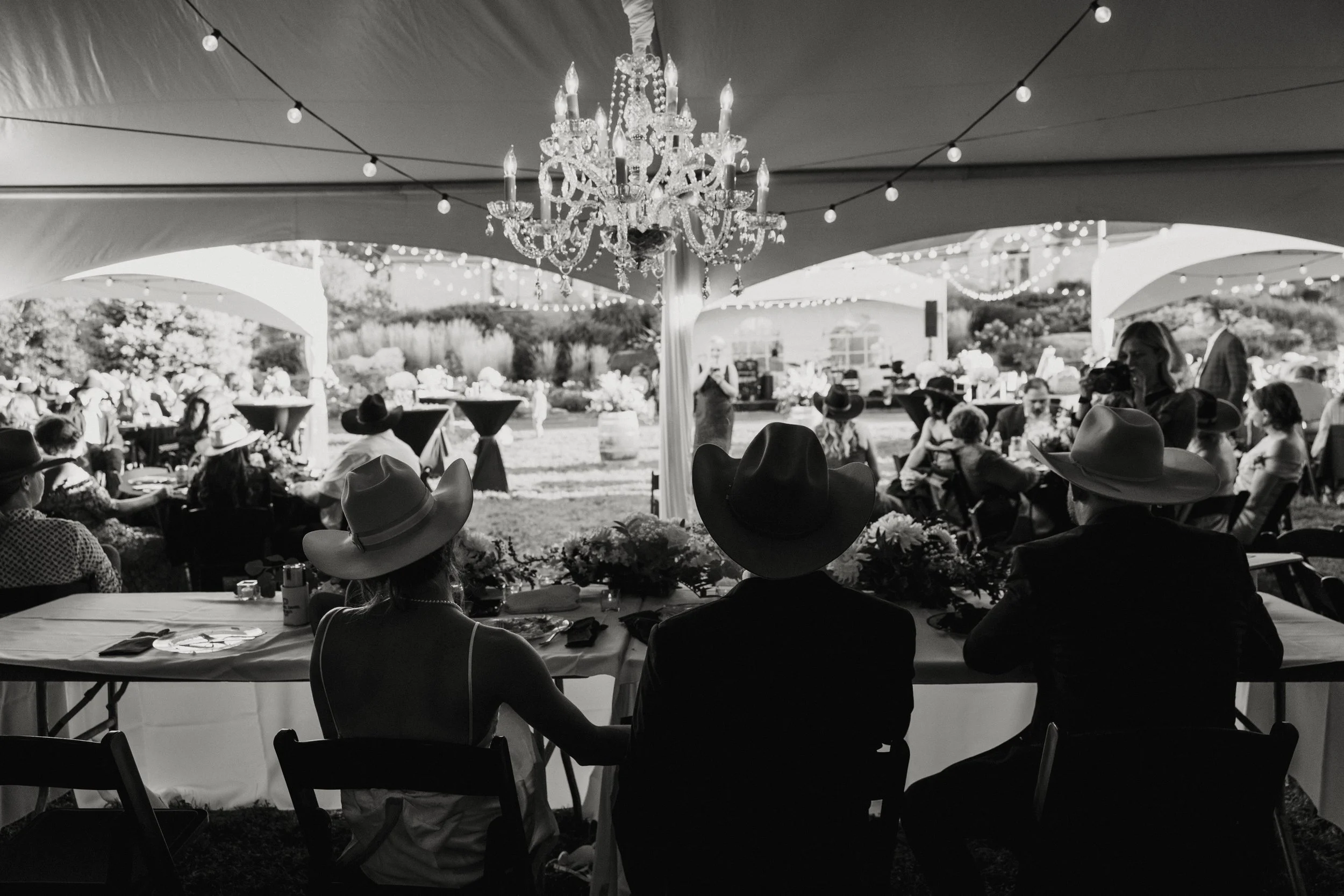 A black and white photo of a wedding reception under a large tent with elegant chandeliers and string lights. Guests wearing cowboy hats are seated at tables, and there is a lush outdoor setting in the background.