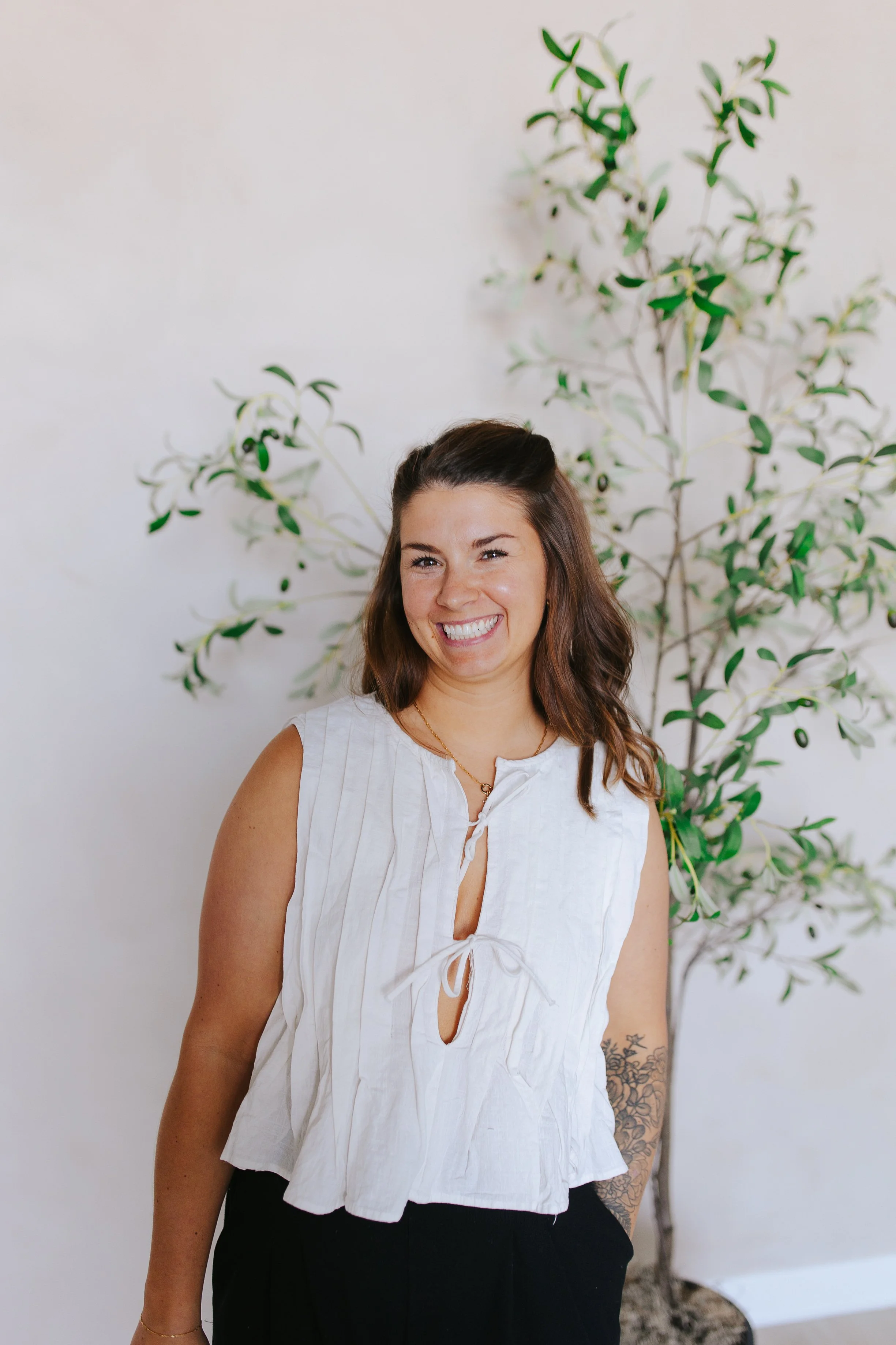 A smiling woman with brown hair in loose waves, wearing a sleeveless white top with tie closures and black pants, standing in front of a white wall with a green leafy plant.