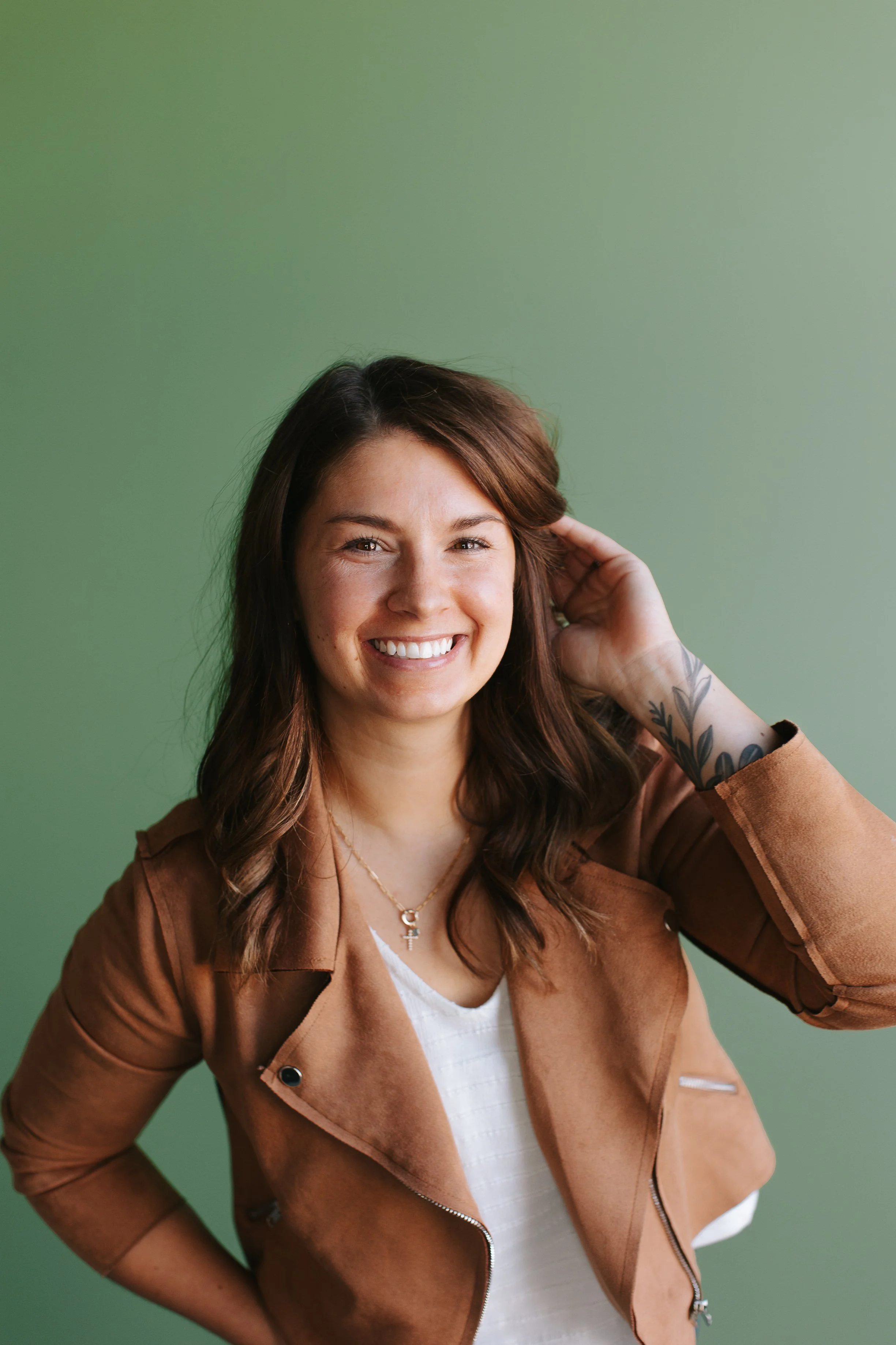 A smiling woman with wavy brown hair, wearing a brown jacket over a white top, standing against a green background.
