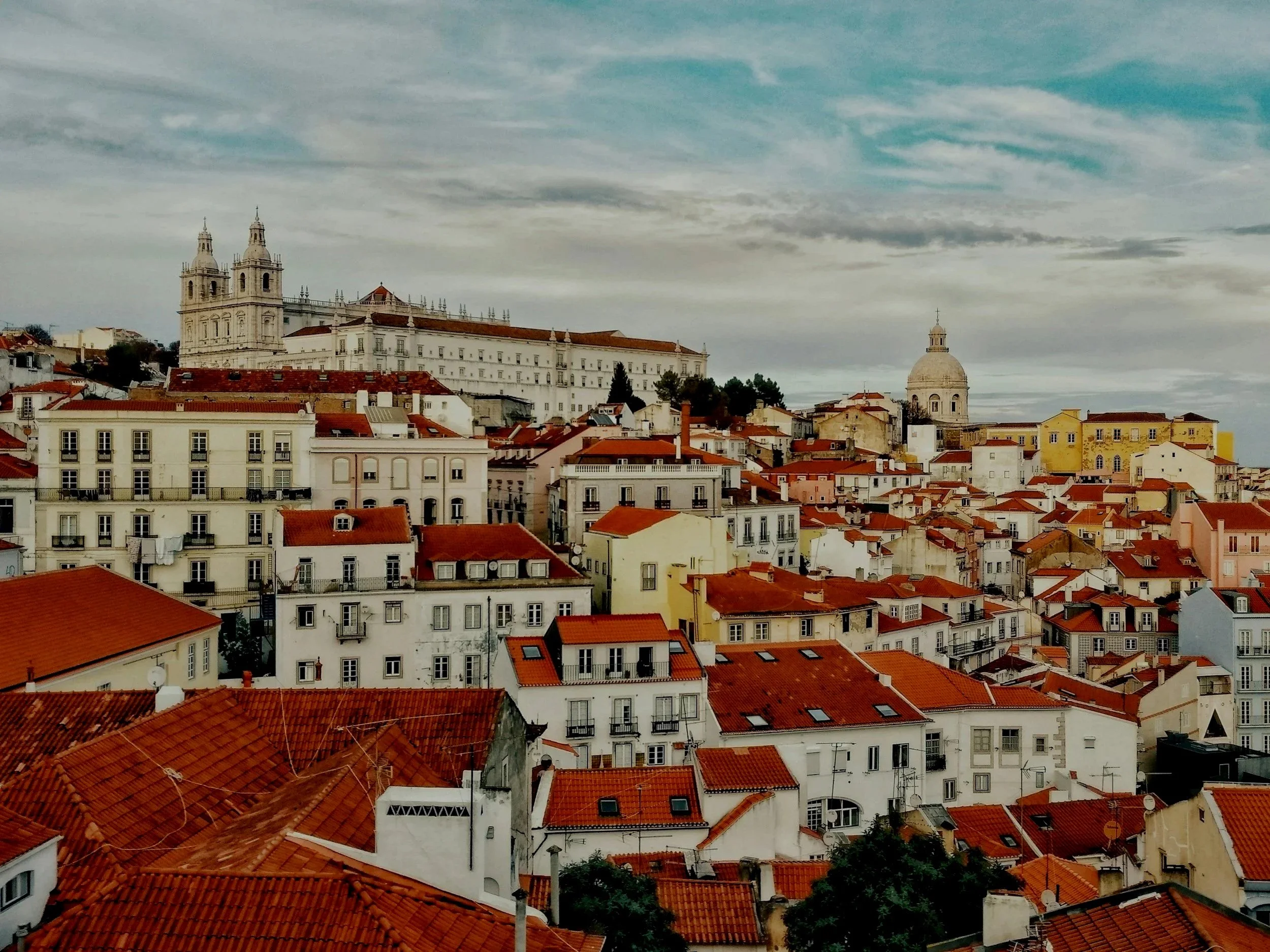 A cityscape of Lisbon, Portugal, with terracotta rooftops, historic buildings, and churches on a partly cloudy sky.