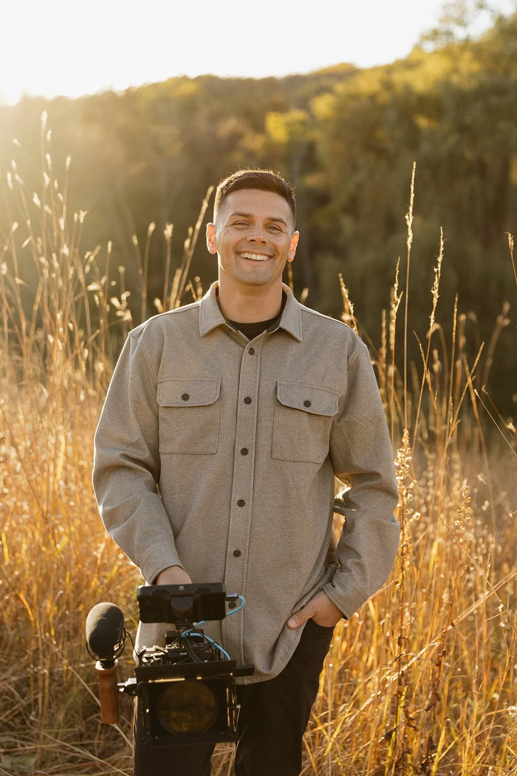 Man smiling in a field holding a video camera, wearing a light gray jacket and dark pants, with sunlit grass and trees in the background.