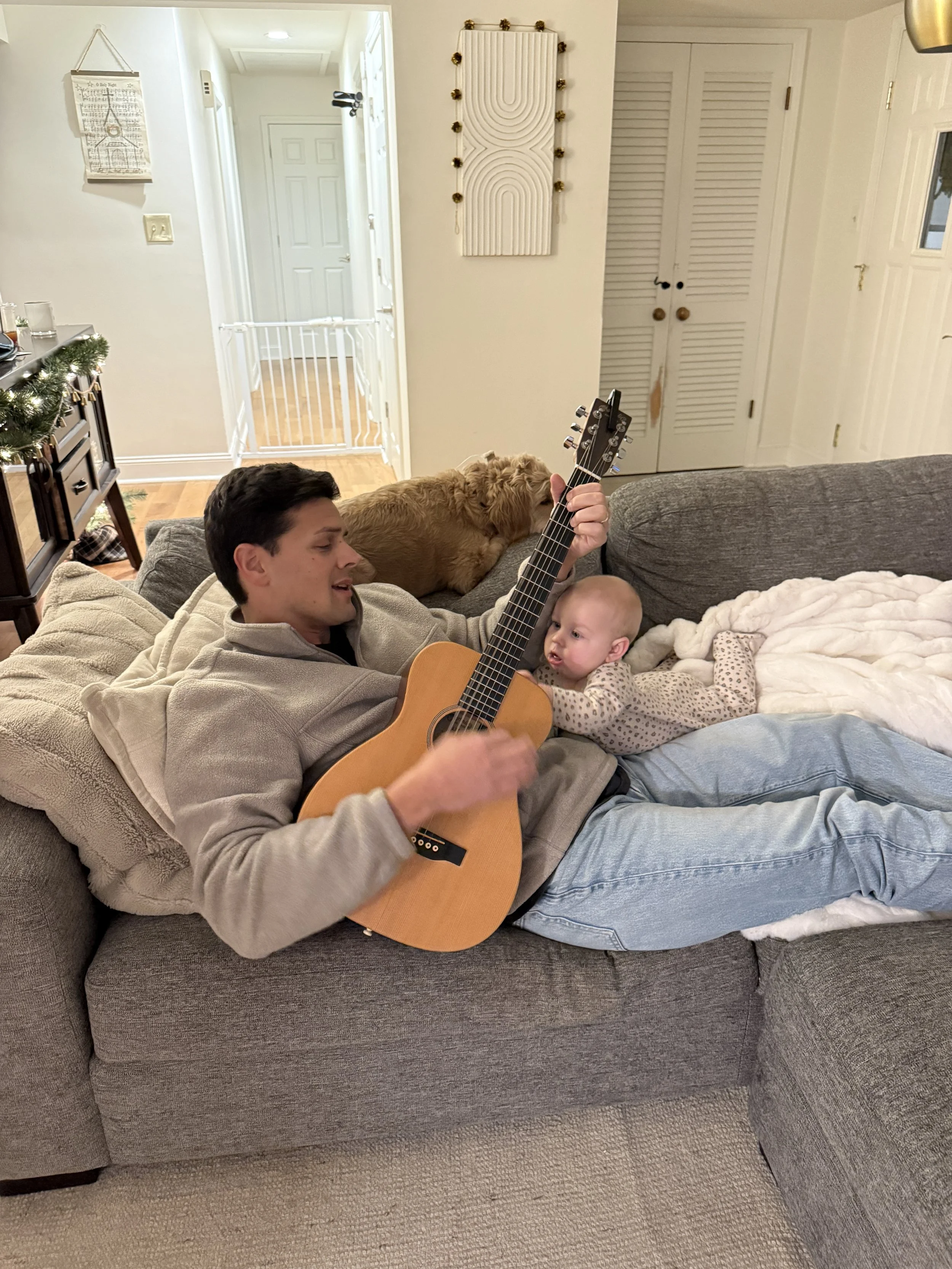 A man lying on a gray couch playing an acoustic guitar while a baby girl lying next to him reaches for the guitar. A small dog is on top of the couch behind them.