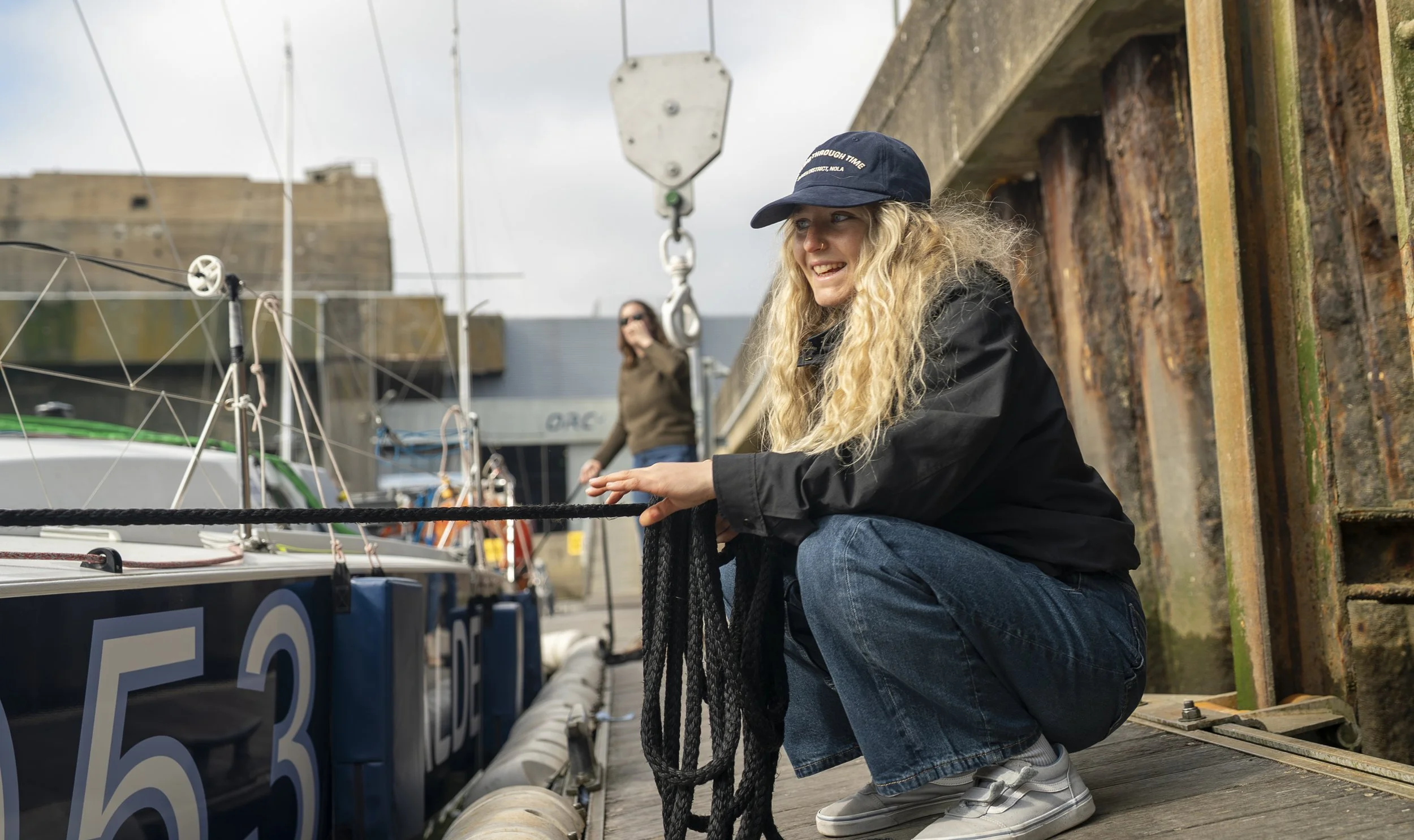 Jeune femme avec une casquette, cerclée à un quai de bateau, tenant une corde, avec un bateau à voile à côté, et un bâtiment en arrière-plan.