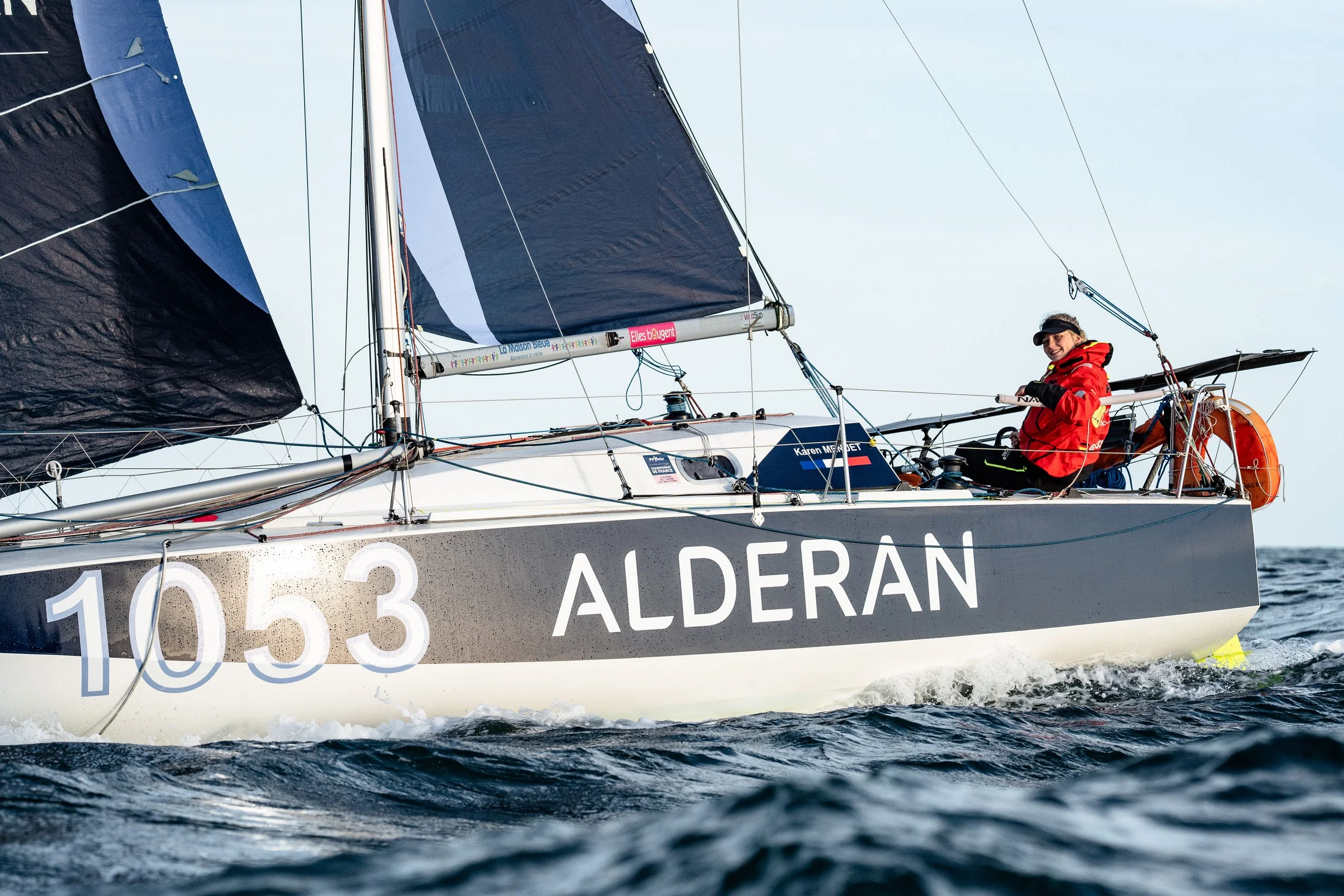 Voilier en mer avec un équipage, la personne à l'arrière porte une veste rouge et un chapeau, le voilier porte le nom Aldéran et le numéro 1053.