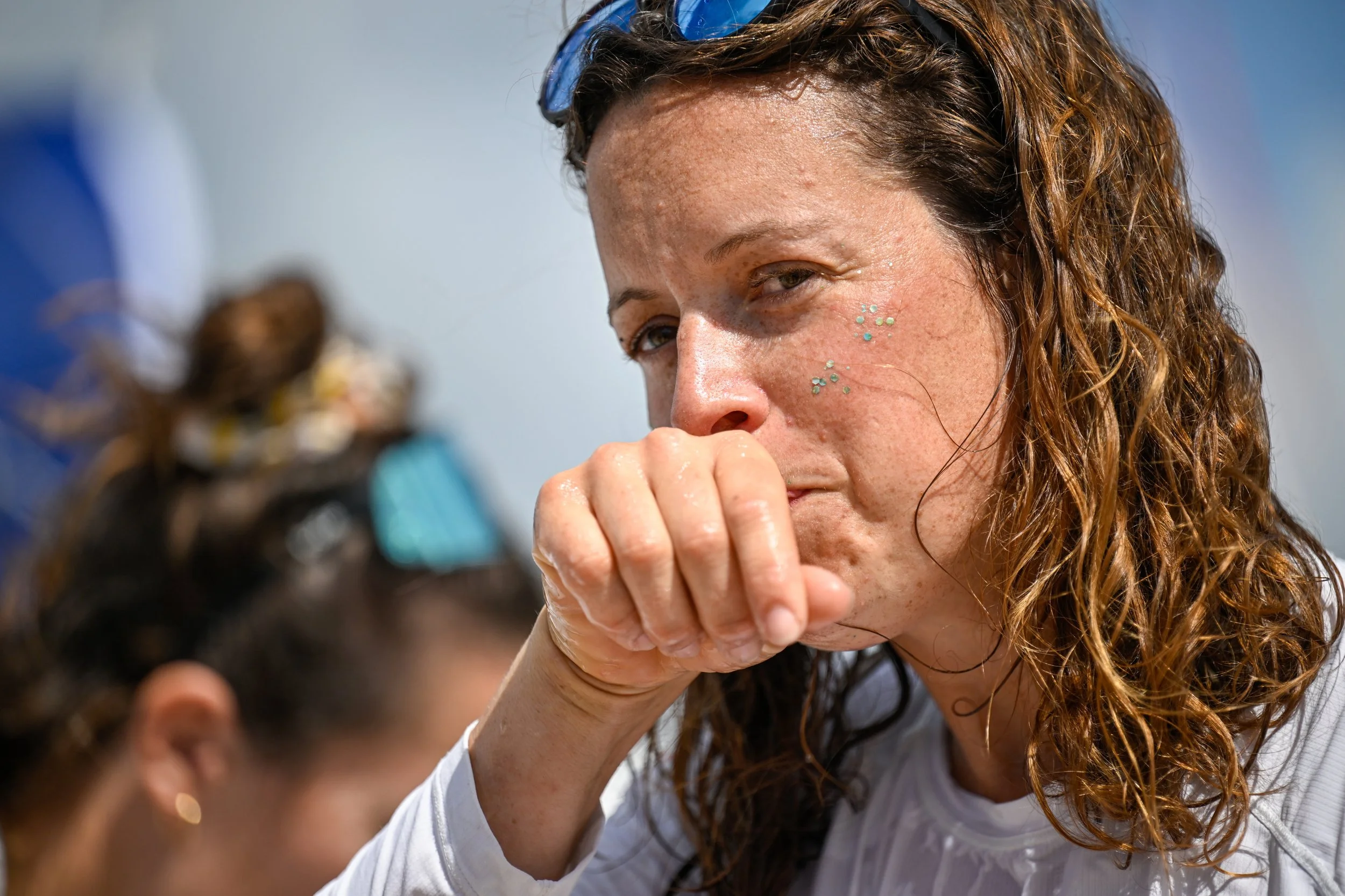 Une femme avec des taches de rousseur et des strass sur le visage, portant des lunettes de soleil sur la tête, semble être triste ou émue, elle couvre sa bouche avec sa main.