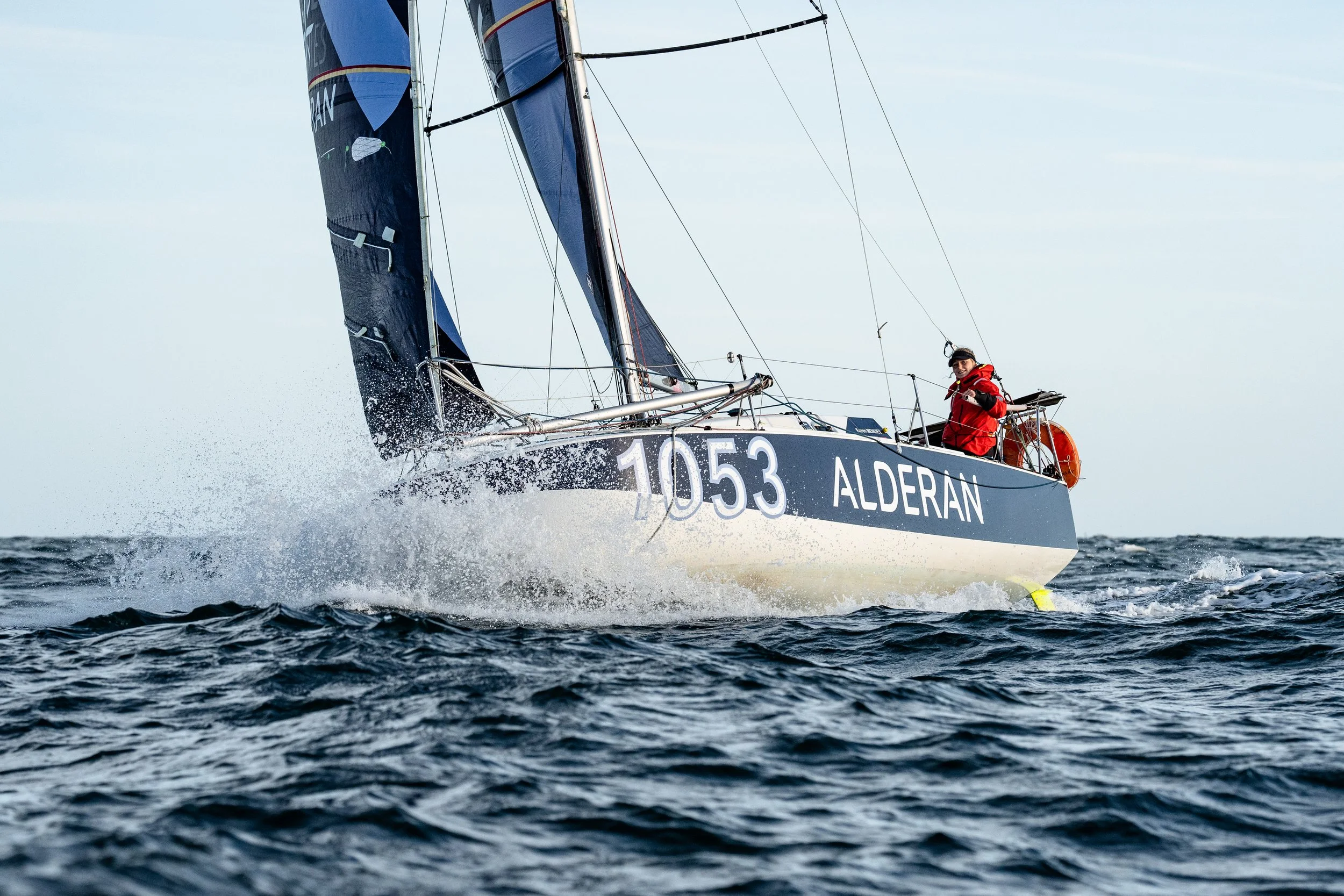 Voilier naviguant sur la mer avec un marin en veste rouge, numéros 1053 et nom Aderan visibles sur la coque.
