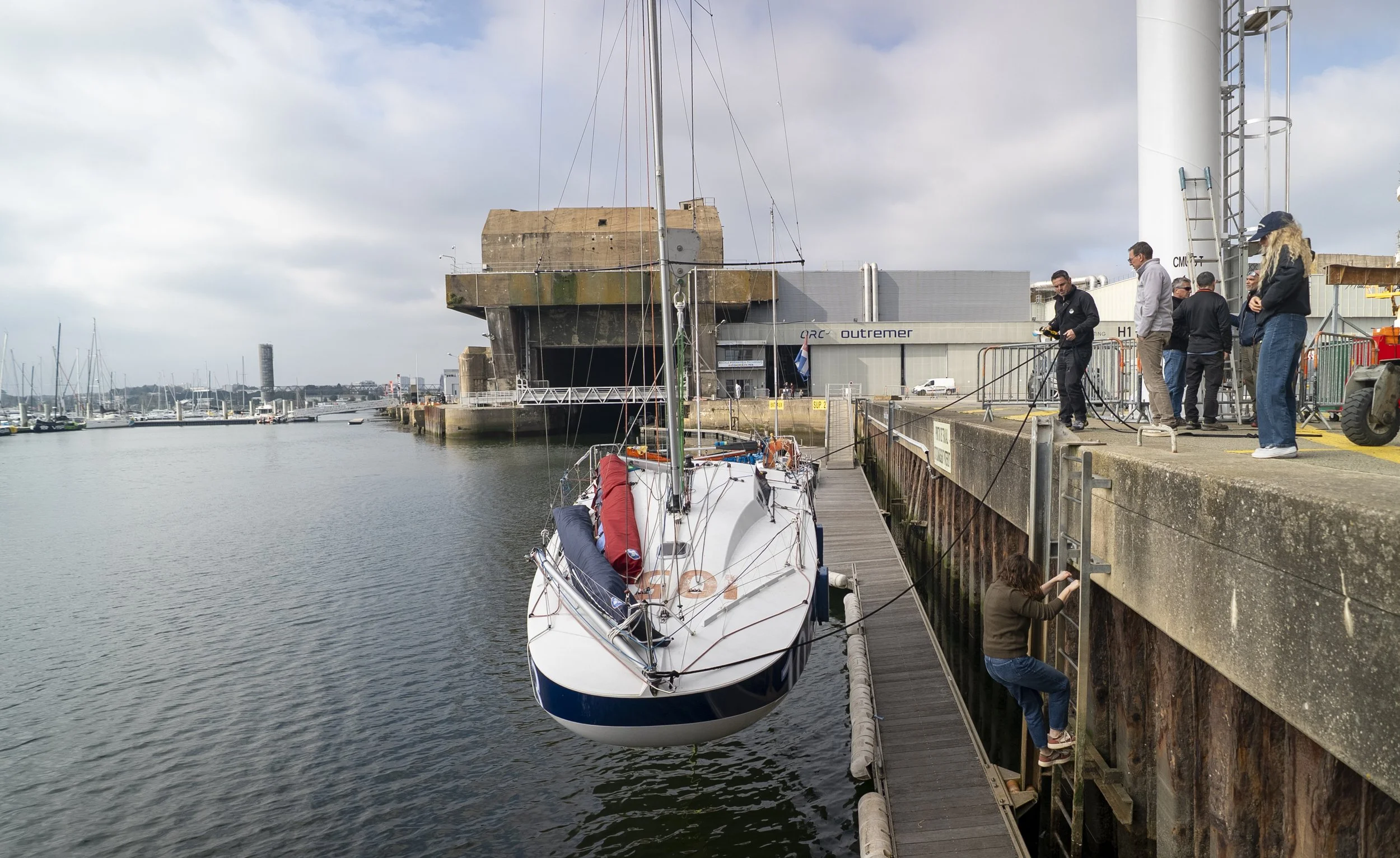 Une femme grimpe sur une échelle le long d'un quai en pierre pour atteindre un bateau amarré, tandis que plusieurs personnes sur le quai prennent des photos ou regardent. Un voilier est amarré dans le port, avec un bâtiment industriel en arrière-plan