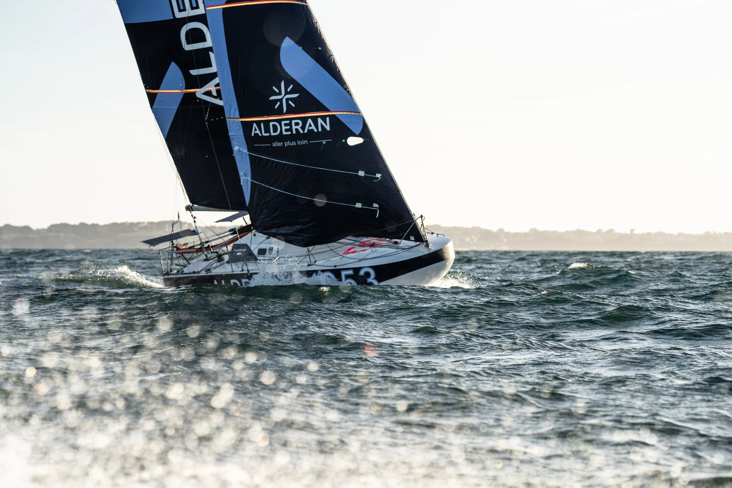 Un voilier en mer avec grande voile noire portant des inscriptions bleues et blanches, naviguant dans des eaux agitées sous un ciel clair, avec une ligne d'horizon au loin.