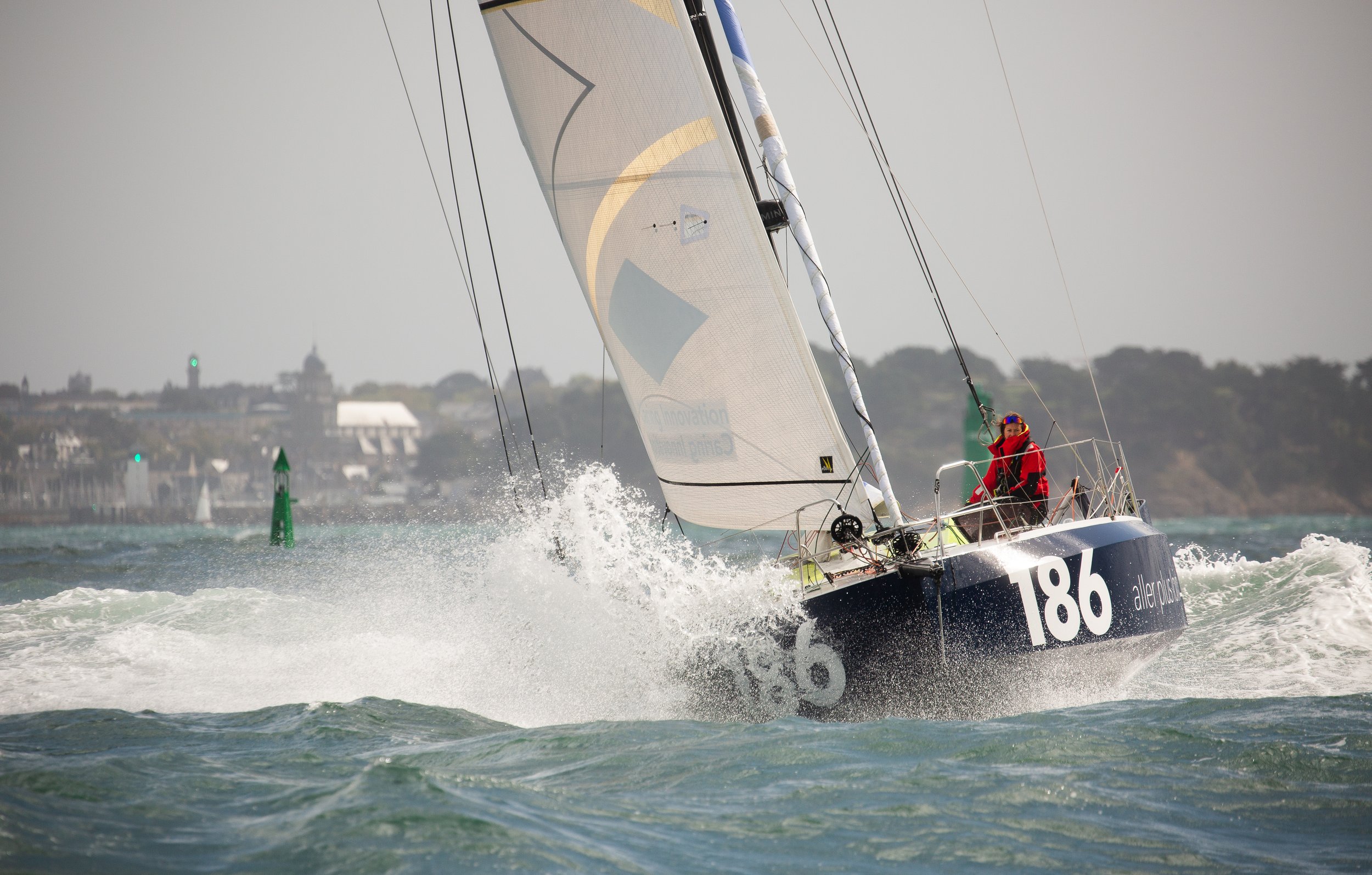Voilier en course avec un skipper portant un gilet rouge naviguant à grande vitesse sur l'eau, avec un fond de ville floue et des marqueurs de navigation
