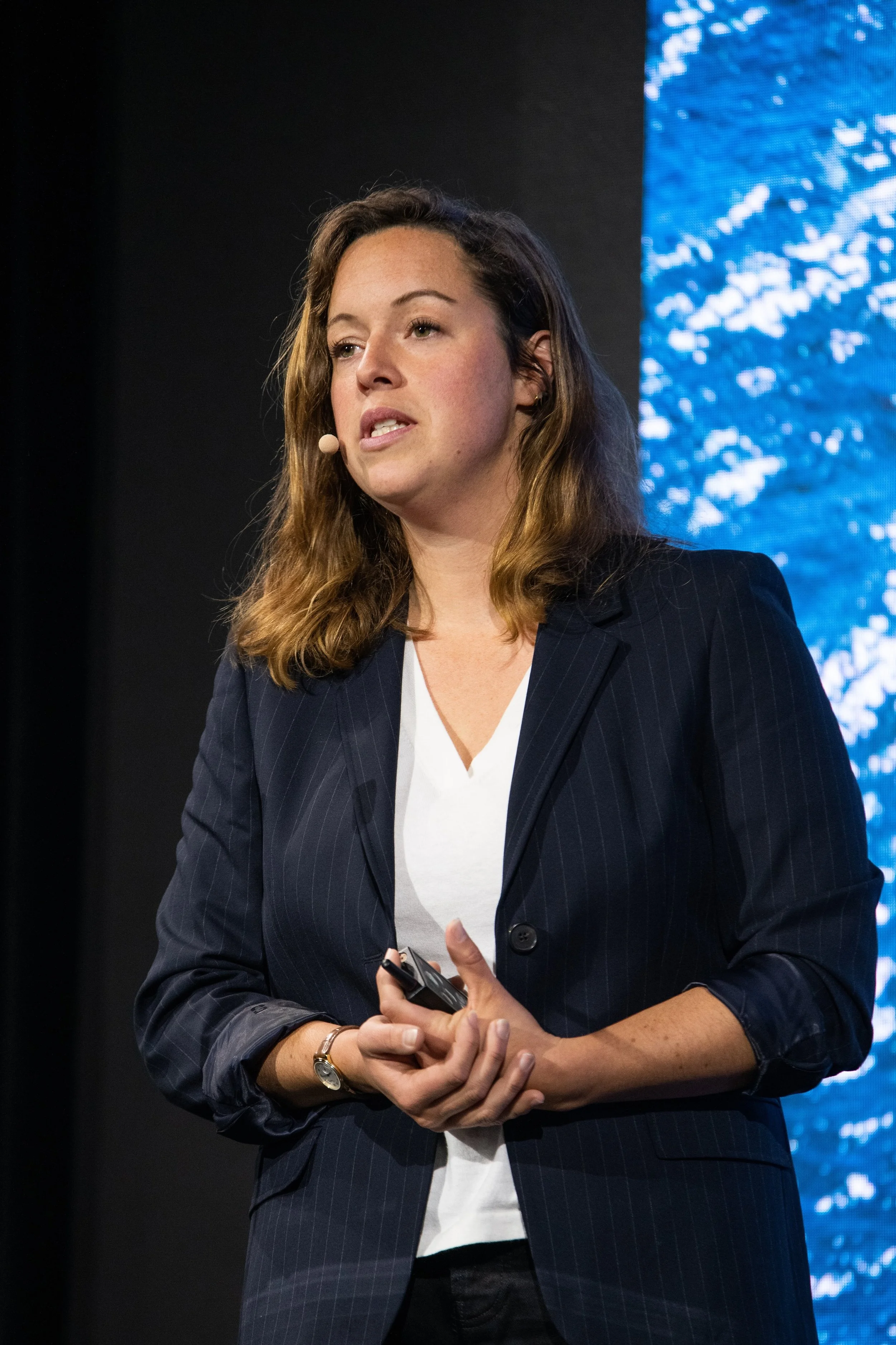 Femme parlant lors d'une conférence, portant une veste noire à rayures, une chemise blanche, et tenant un dispositif de contrôle, avec un décor numérique en arrière-plan.