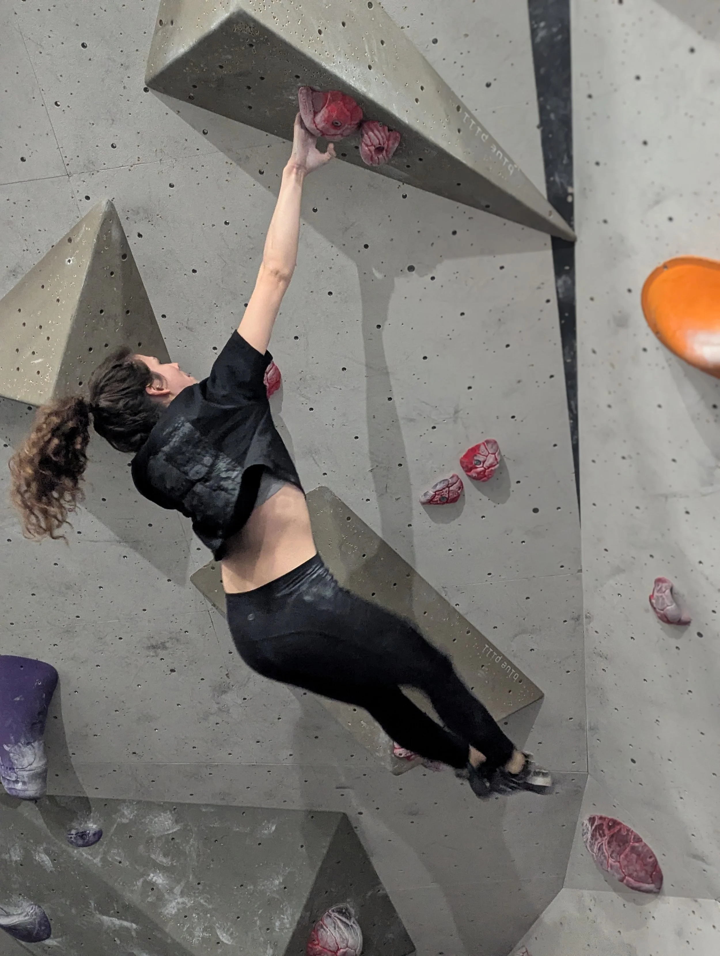 A climber wearing a T-shirt and loose-fitting trousers on an indoor bouldering wall. Climbing clothes don't need to be complicated — freedom of movement is the important bit.