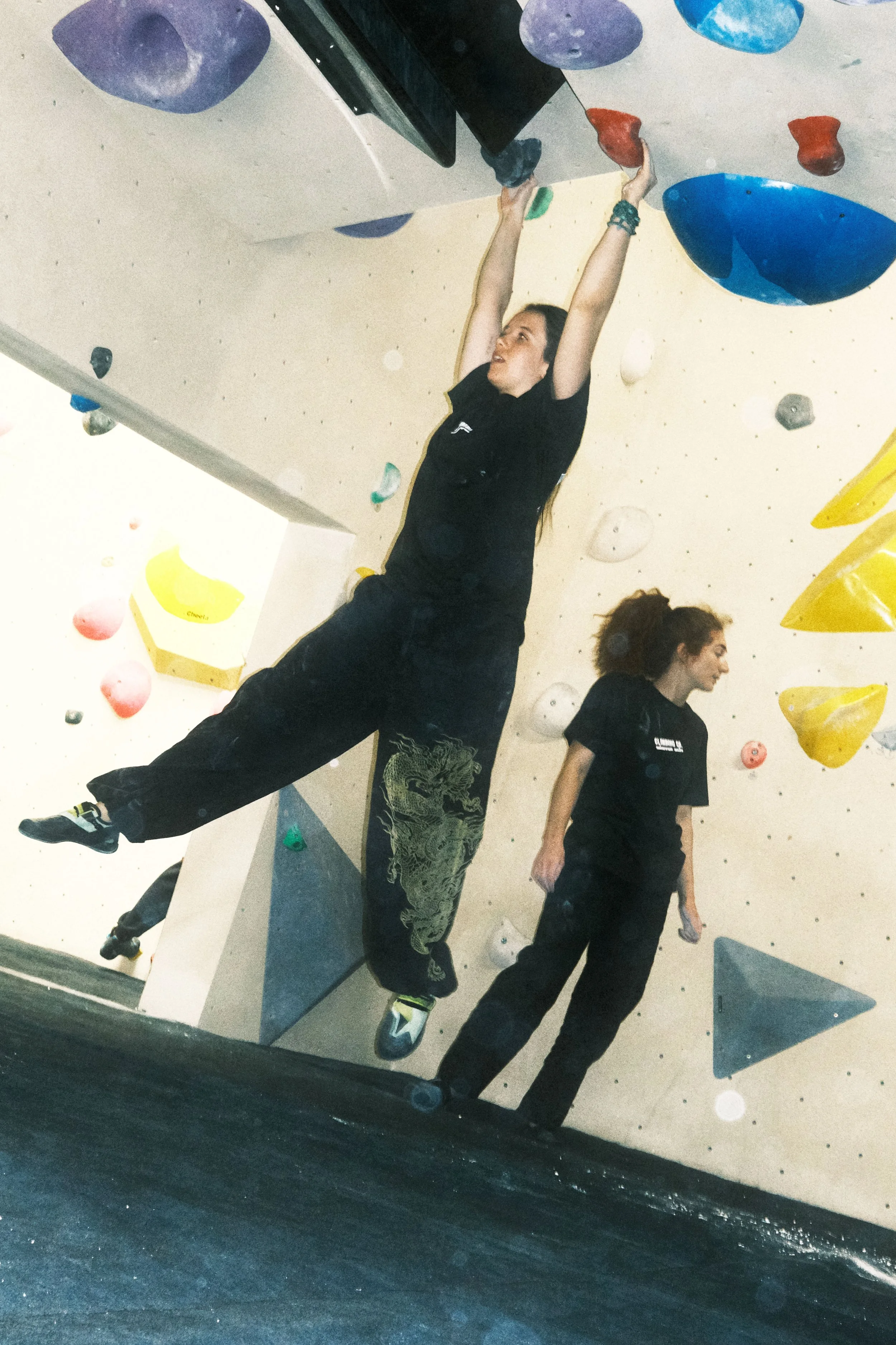 Indoor climber wearing a black Winter Grit x Climbing Co T-shirt while reaching across a bouldering wall.