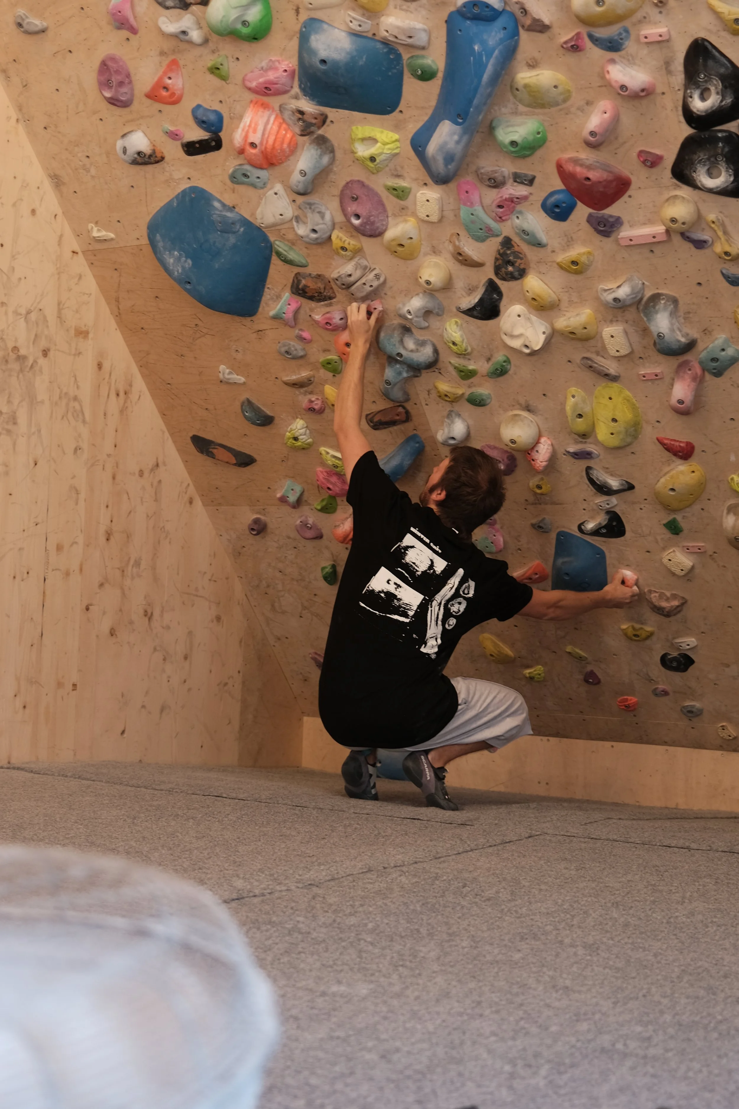 Climber wearing a black Winter Grit Rest Day Volume 2 Tee while bouldering on a steep indoor climbing wall with colourful holds.
