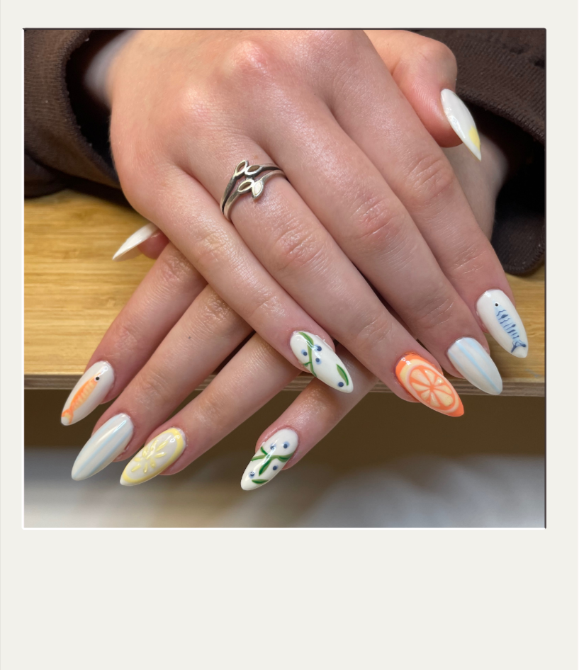Hands with colorful, floral and fruit-themed nail art, resting on a wooden surface, wearing a silver ring with leaves.