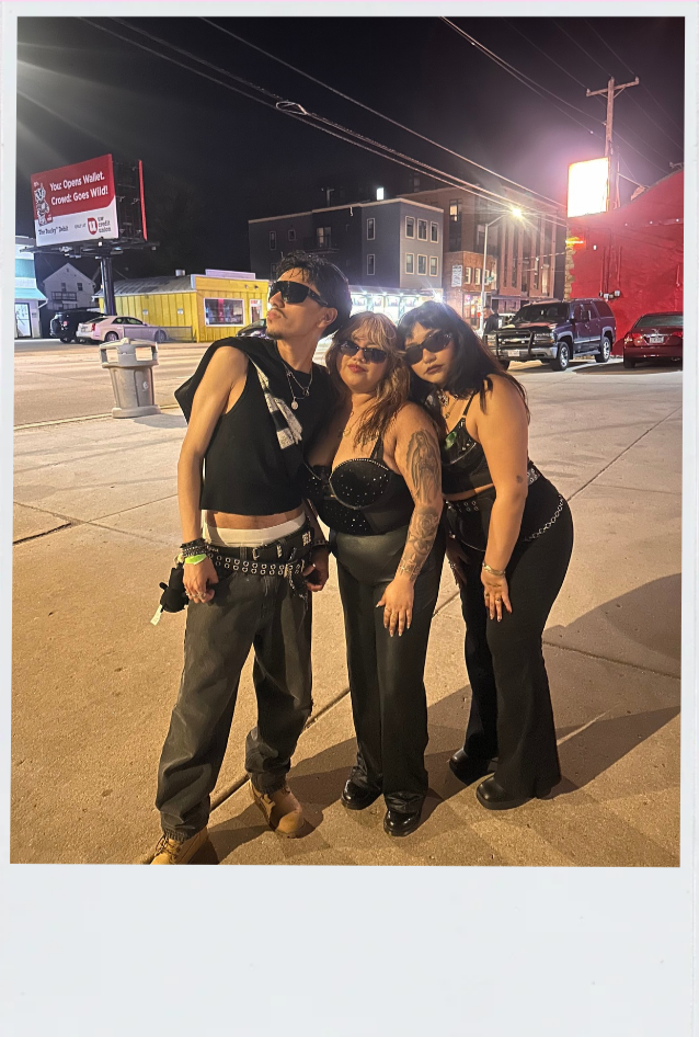 Three women standing together on a city street at night, wearing dark clothing and sunglasses.