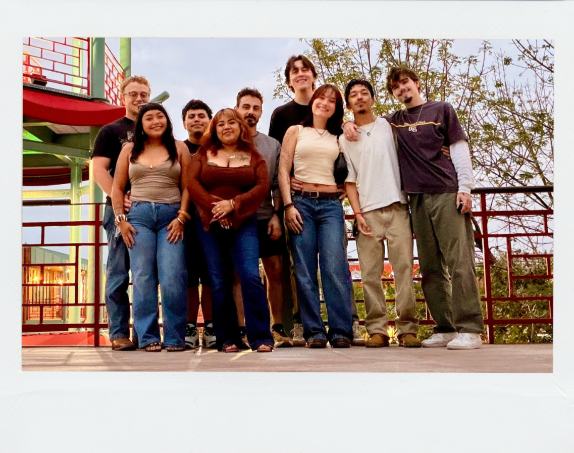 Group of nine diverse young adults standing together outdoors, smiling, on a wooden platform with trees and a colorful playground structure in the background.