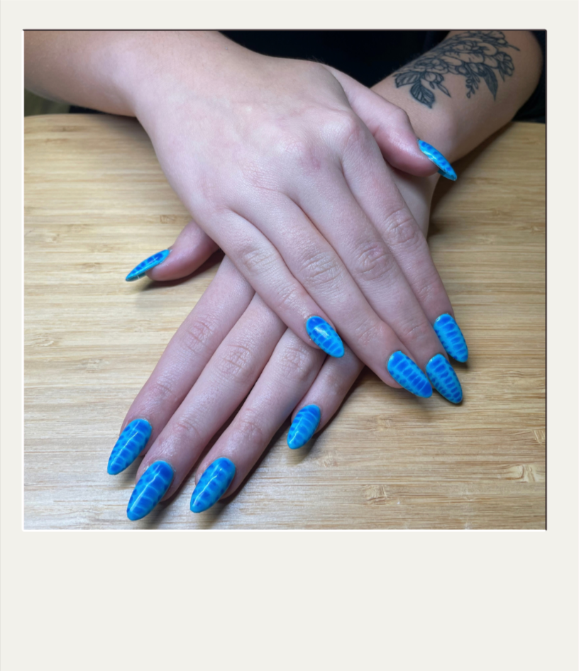 A person's hands resting on a wooden surface, with blue and black patterned nail polish, and a tattoo on one wrist.