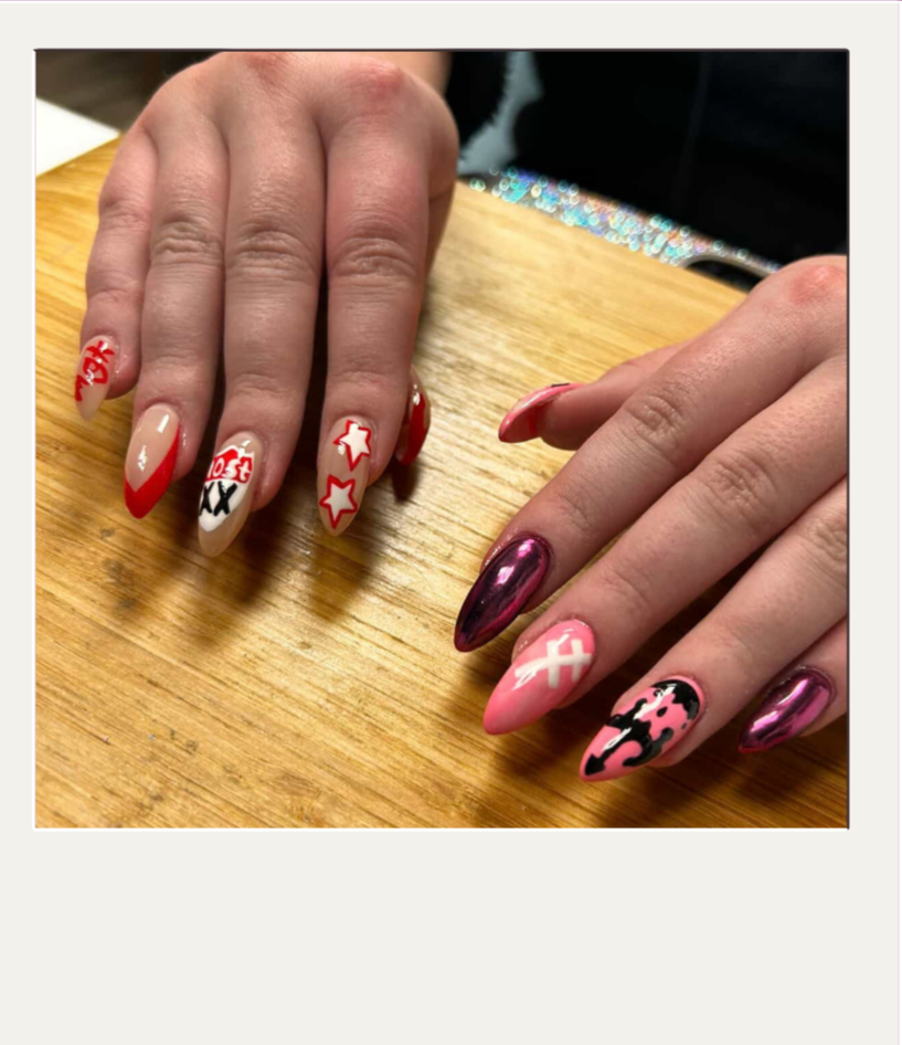 Close-up of two hands with decorated nails resting on a wooden surface, featuring red, pink, black, and white nail art with various designs including stars, a cross, and animal print patterns.