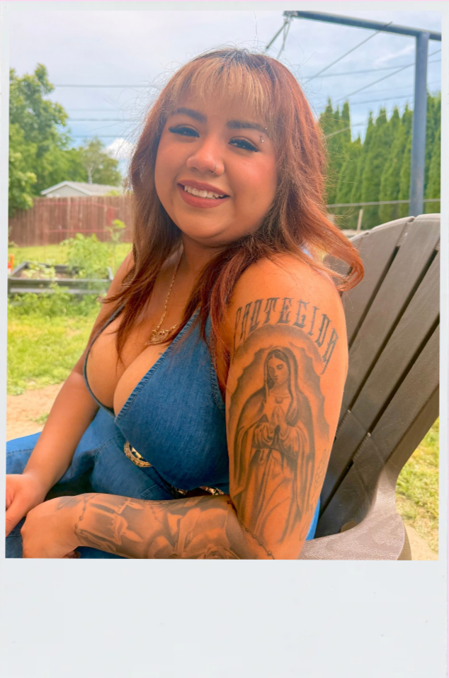 Young woman with tattoos of religious figures and text on her right arm, smiling outdoors on a wooden chair.