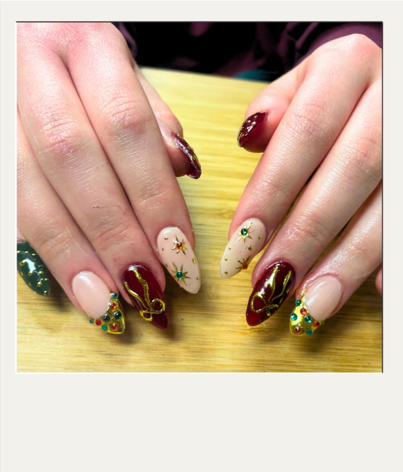 Close-up of hands with decorated Christmas-themed nail art featuring red, green, and gold designs, including bows, stars, and rhinestones, on a wooden surface.