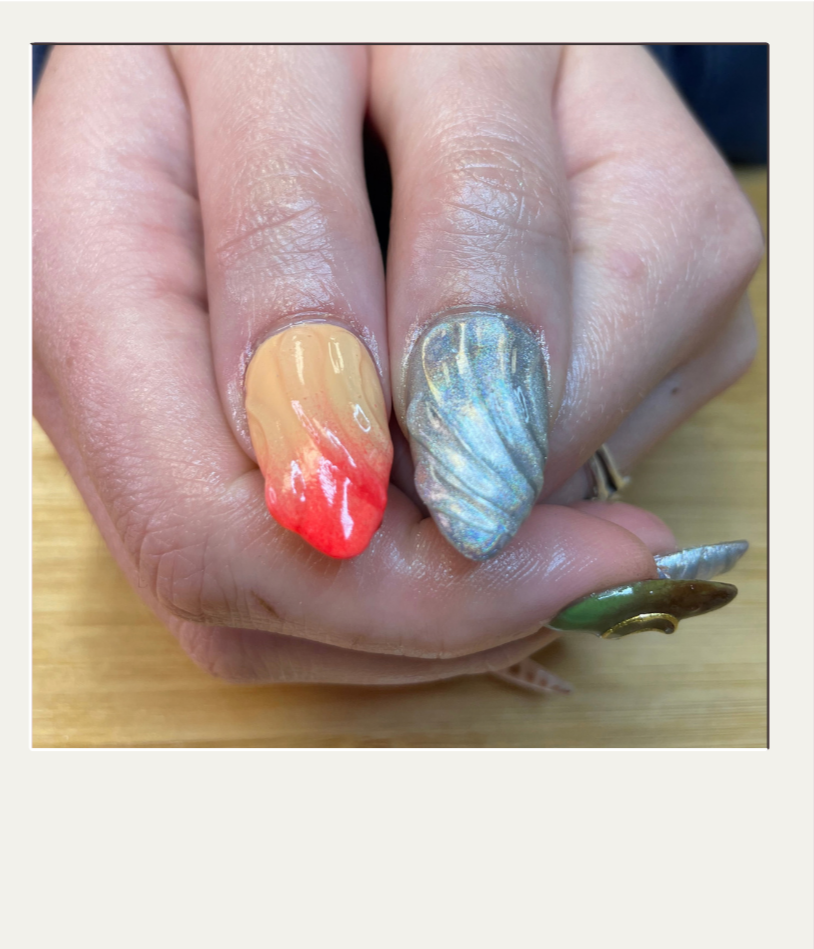 Close-up of a hand with creatively painted and decorated fingernails. The thumbnail has three fingers visible, each with different nail art designs: one with a red and beige gradient, one with a metallic blue swirl, and the thumb painted green with gold accents.
