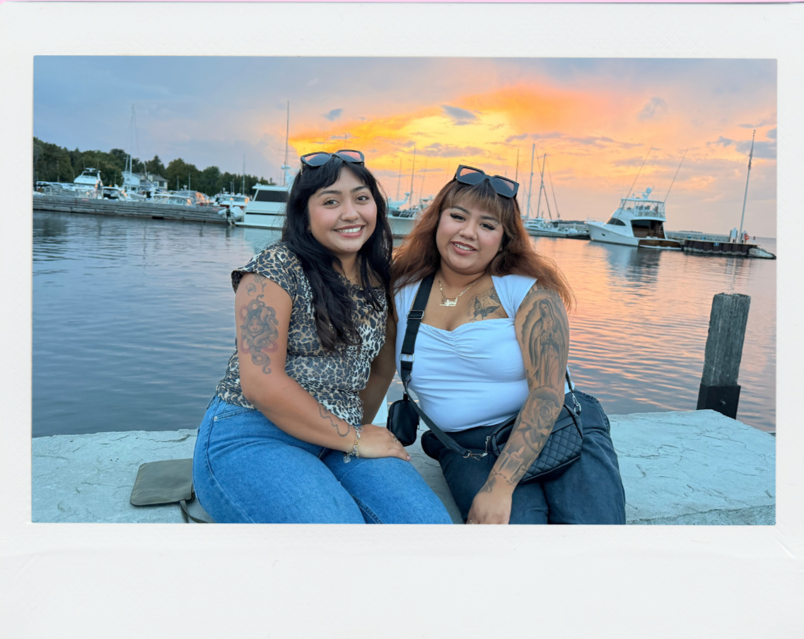 Two women sitting on a dock near the water with boats and a sunset background.