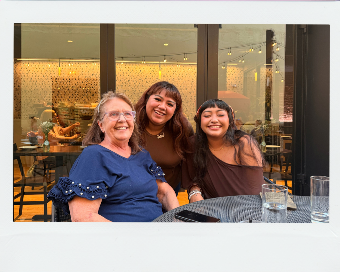Three women smiling and sitting at a table in a restaurant or cafe with glass walls and warm lighting.
