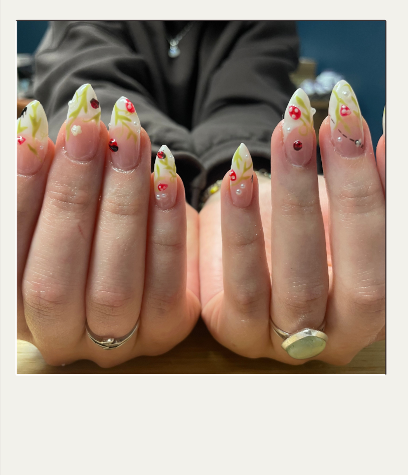 Close-up of hands with decorated nails, featuring a floral and ladybug design, wearing rings, with a person in a dark jacket in the background.