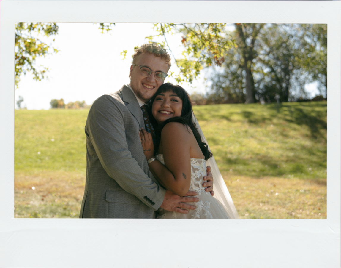 A smiling couple, dressed in wedding attire, hugging outdoors on a sunny day with trees and grass in the background.