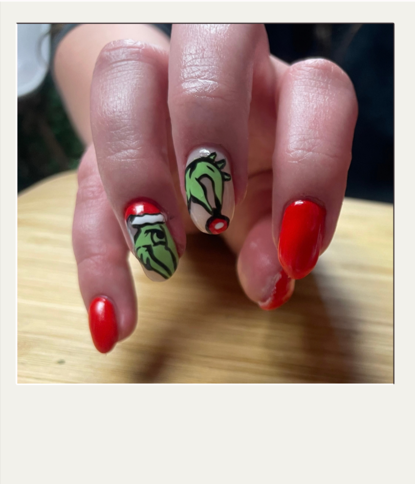 Close-up of a hand with red-painted nails featuring white flower designs, wearing a ring with a white stone, against a background of a living room with a lamp and a television, christmas nails