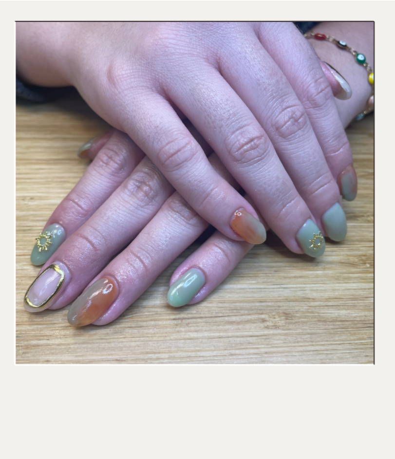 Close-up of hands with pastel-colored manicured nails featuring decorative elements like sun symbols and gold accents, resting on a wooden surface.