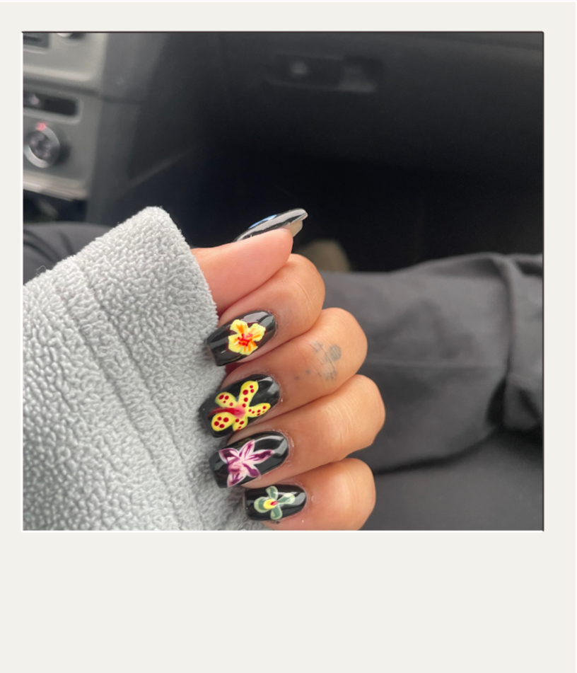 Close-up of a hand with black nail polish featuring colorful floral and butterfly designs, held in a gray fleece glove inside a vehicle.