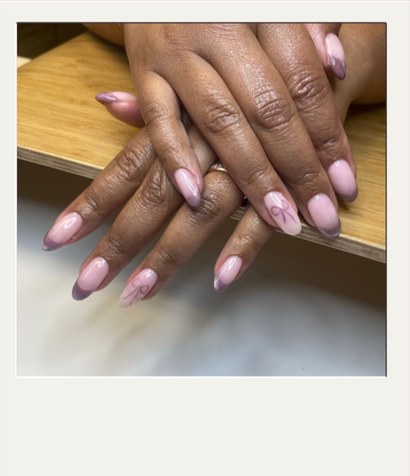 Close-up of two hands with manicured nails featuring pink and purple ombre style and small ribbon designs, resting on a wooden surface.