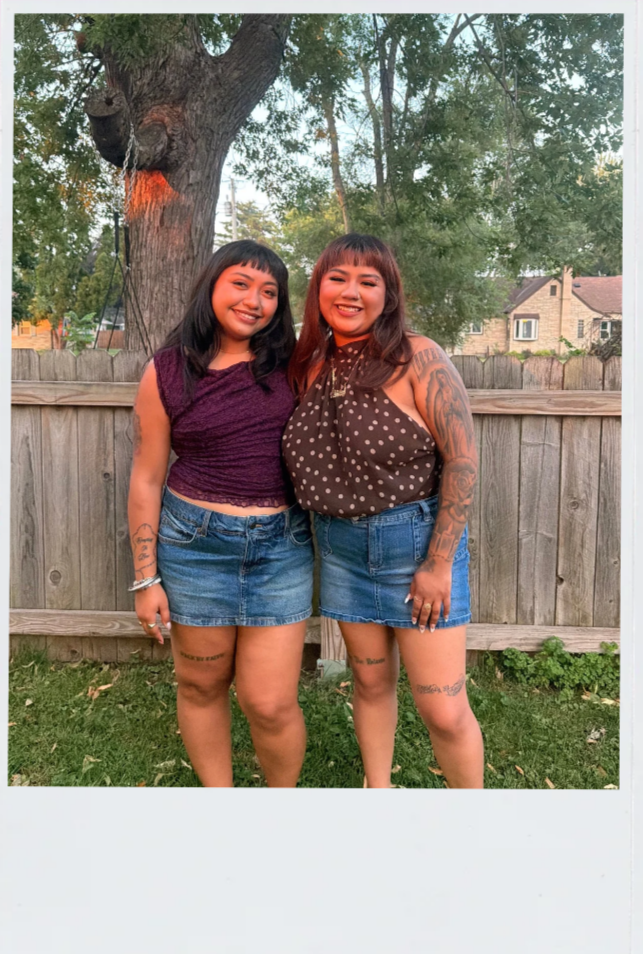 Two women standing outside in front of a wooden fence and a tree, smiling and posing for a photo. Both are wearing denim skirts and sleeveless tops, with visible tattoos on their arms and legs.