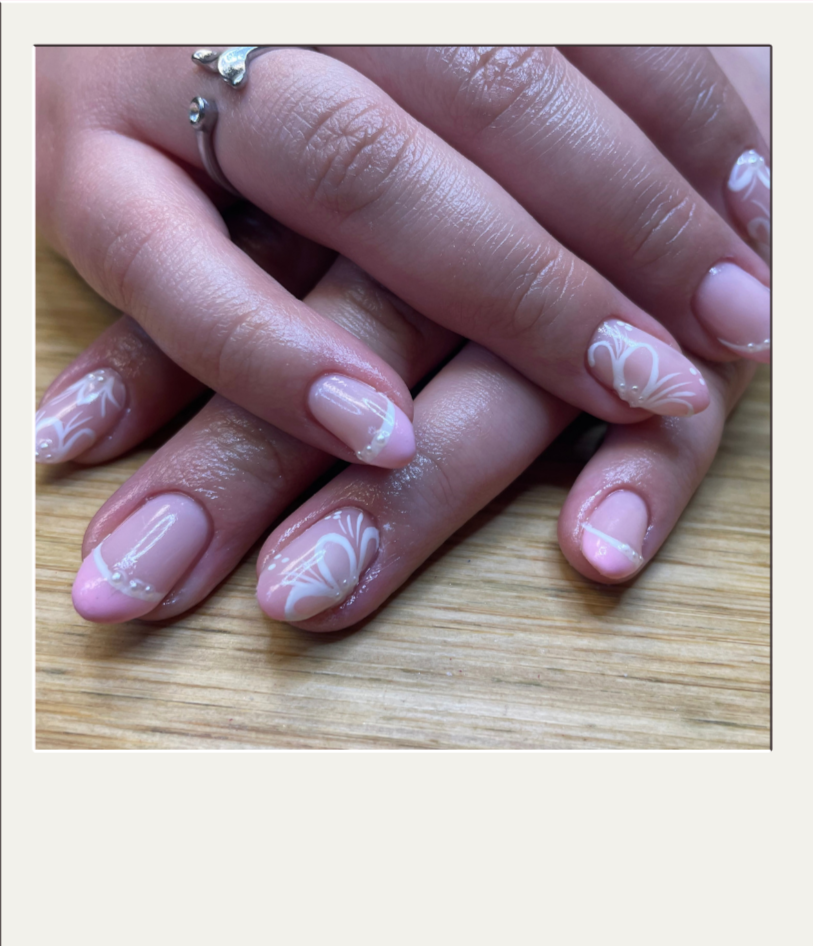 Close-up of hands with manicured nails featuring pink and white French tip nail art with delicate white swirl designs on a wooden surface.