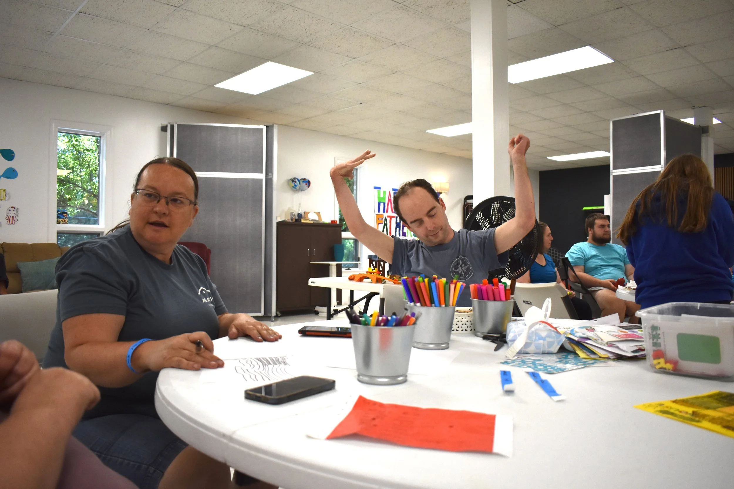 A group of people are gathered around a large, white circular table in a brightly lit community room or classroom, engaged in an activity. In the foreground on the left, a woman wearing glasses and a dark grey t-shirt sits at the table, looking towar