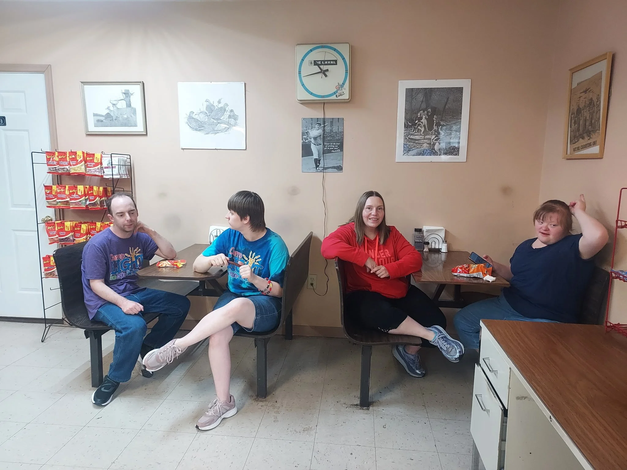 Alt Text: Four people sit at two separate small wooden booths inside a casual dining area or breakroom with light beige walls. On the left, a man in a purple t-shirt and jeans sits across from a person wearing a blue tie-dye shirt and denim shorts wh